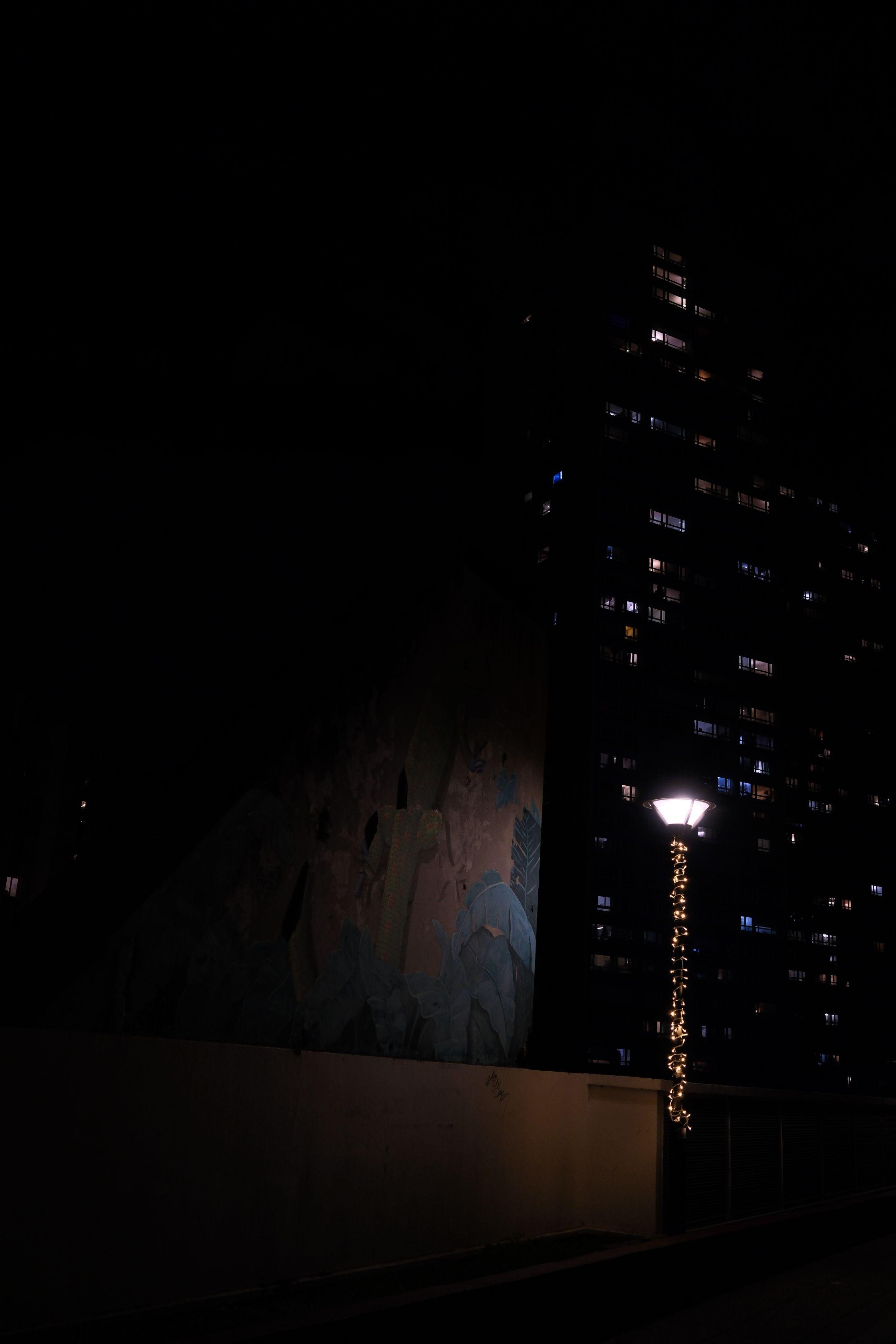 Photo verticale de nuit. Un petit lampadaire dont le poteau est décoré d’une guirlande LED, éclaire timidement un mur qui est couvert de street art rose et bleu