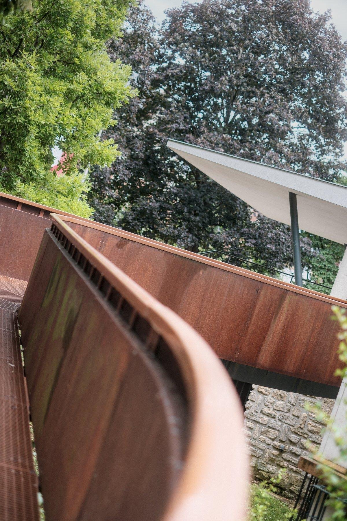 Photo verticale. Vue à ras de rampe d’une passerelle piétonne sinueuse en métal couleur rouille, derrière on voit des arbres verts et marron presque noir, et le toit blanc d’un préau