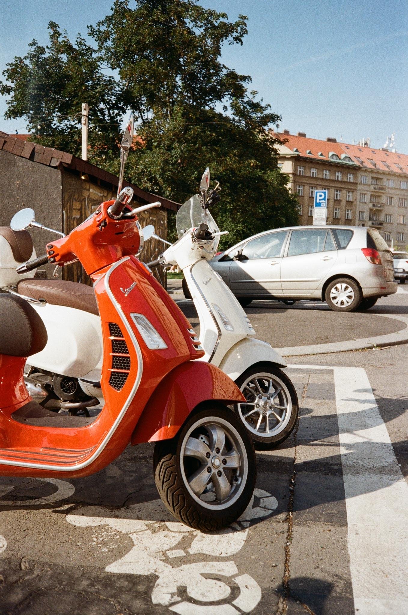 Red and white scooters parked at the curb on a marked lane, with a silver car passing on the street; trees and an apartment building with a red roof in the background under a clear sky.