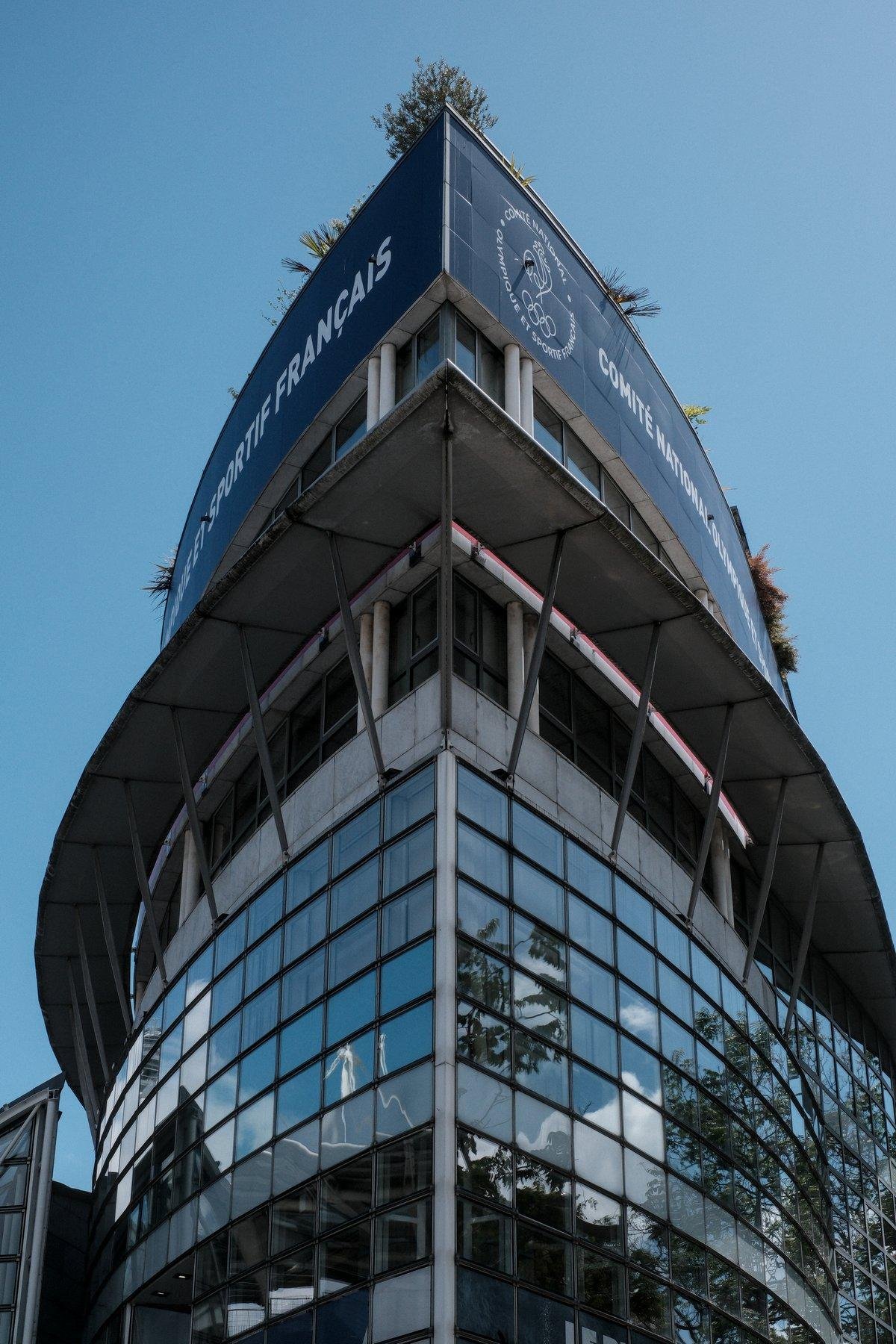 Photo verticale. Contre plongée sur la pointe d’un bâtiment moderne majoritairement en verre/acier et un peu de béton, qui se fini comme la proue d’un bateau