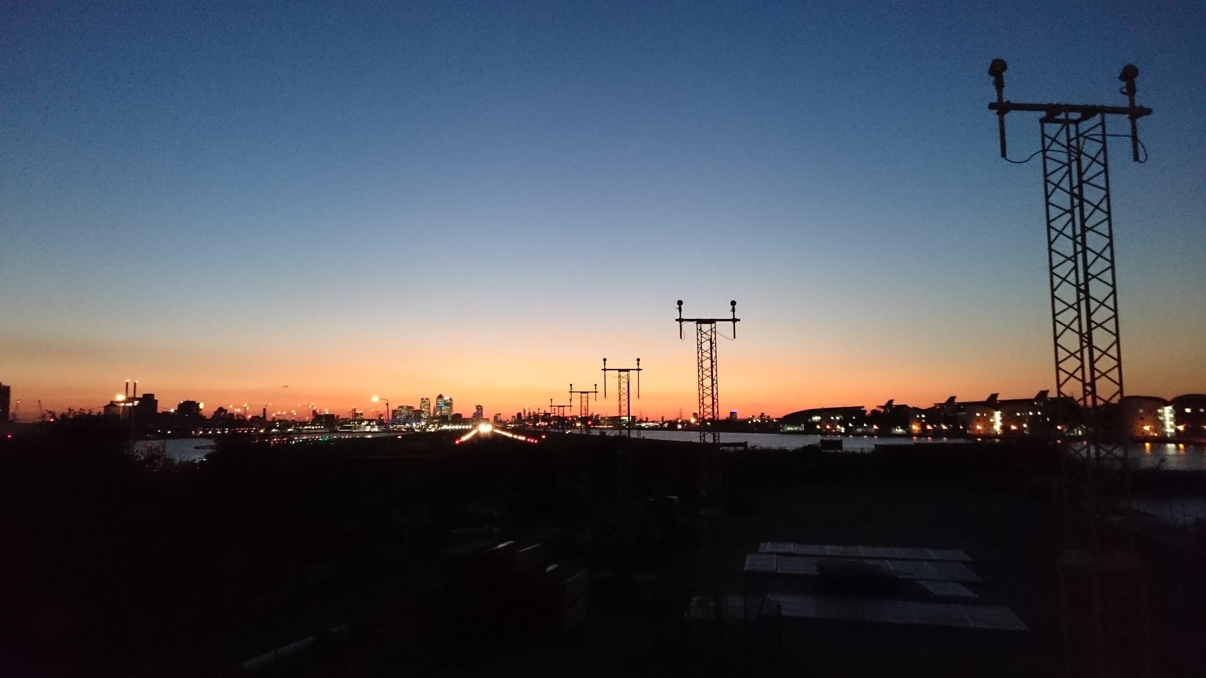 The image shows an airport runway during sunset or sunrise. The sky is a gradient of colors ranging from dark blue to orange and yellow near the horizon. There are silhouette-like structures, possibly runway lights or towers, and the city skyline is visible in the background. The runway lights are illuminated, creating a leading line effect towards the horizon.