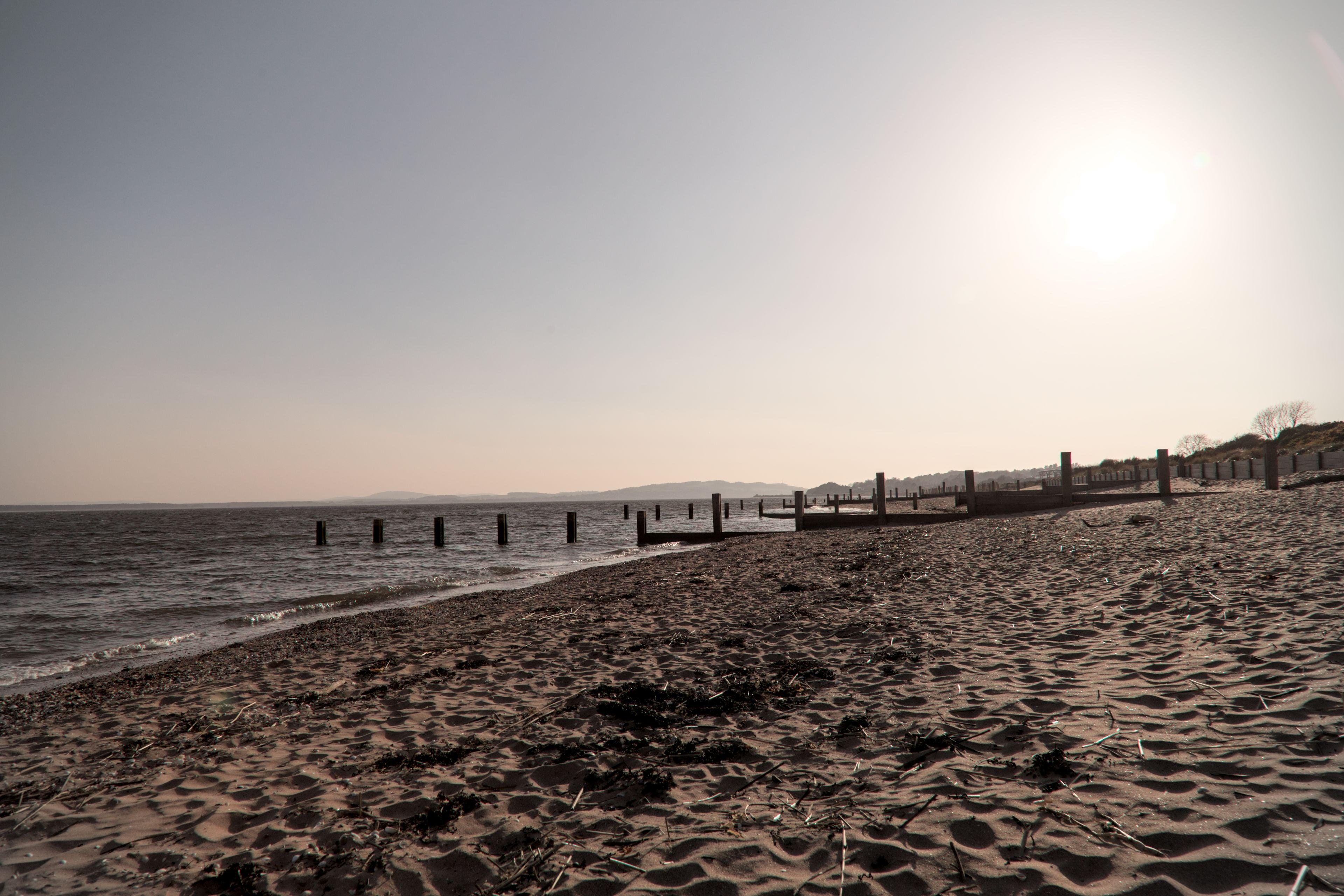 Photo horizontale. Une plage de sable, avec des plot en bois enfoncés dans le sol en ligne vers la mer, sous un ciel gris avec un soleil tout pâle et brumeux