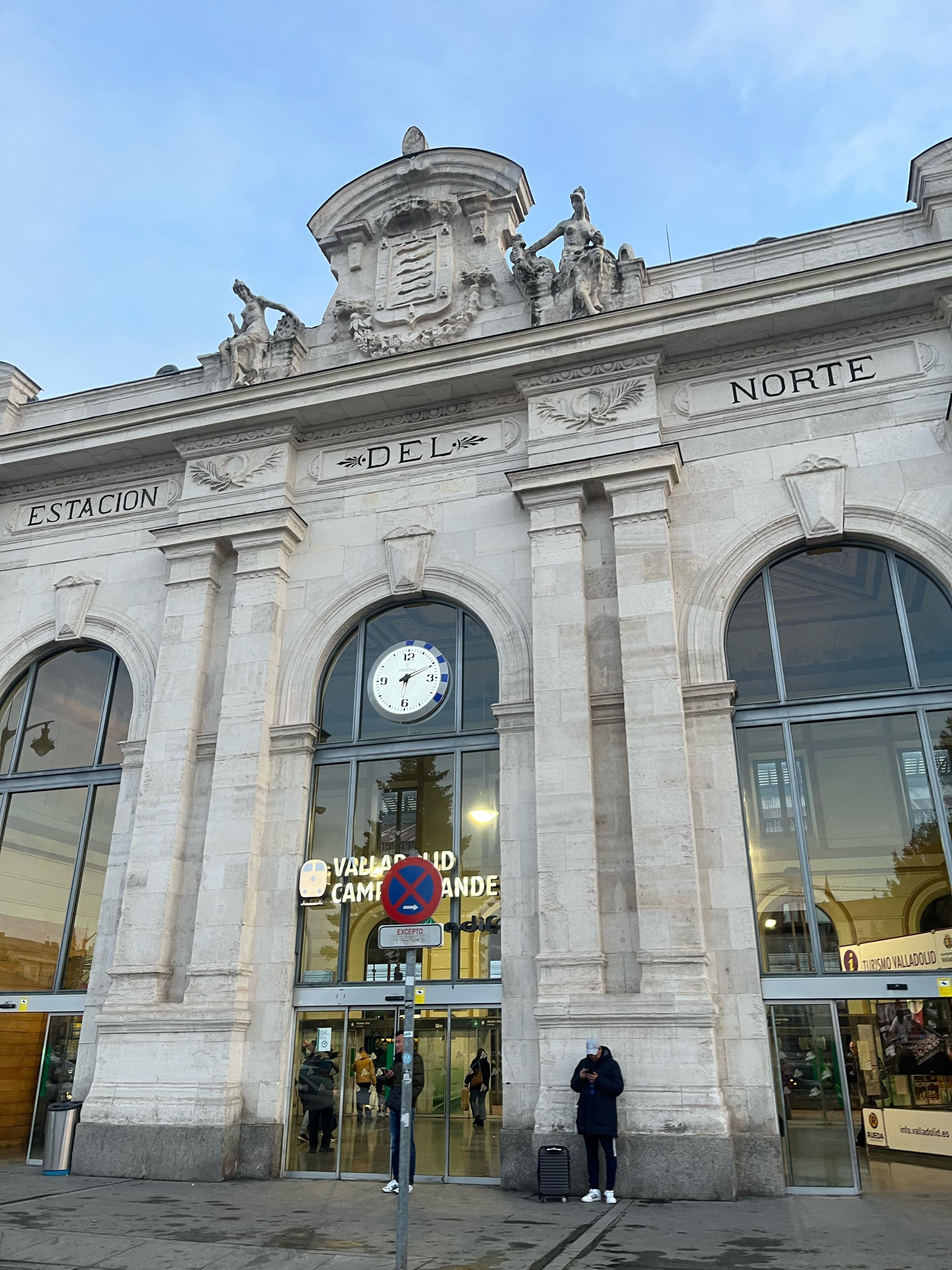 This image shows the facade of a historic building labeled "ESTACION DEL NORTE." It features large arched windows, a clock above the entrance, and sculptures on top. The location appears to be a transportation hub, as indicated by the people and signs around the entrance.