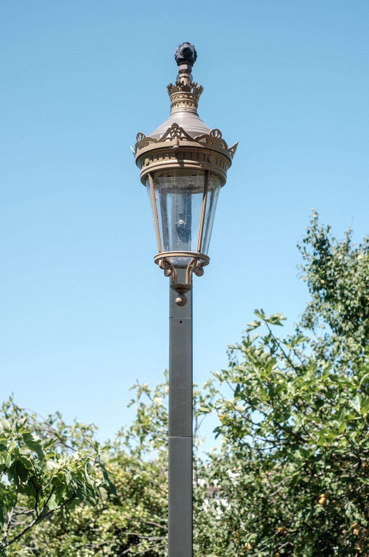 Photo verticale. Vue sur un lampadaire à la lampe très décoré mais au poteau totalement carré sans artifice, le tout vu de face, la lampe alignée avec le poteau, sur fond de ciel bleu en haut et de végétation verte en bas