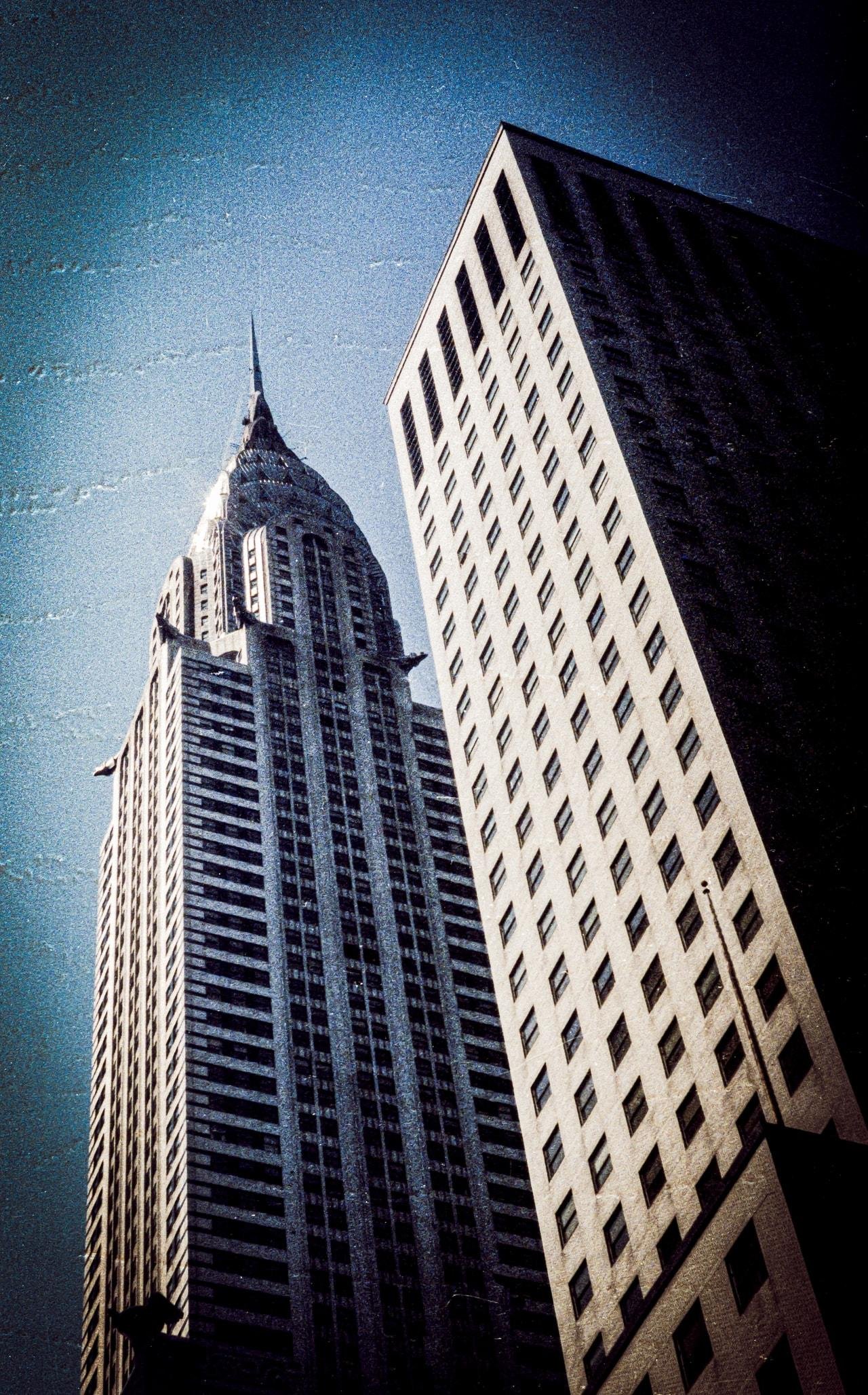 Two skyscrapers rising against a clear blue sky, with the left building featuring an ornate spire and art deco design, while the right building has a simple rectangular shape with multiple windows. The perspective is upwards, capturing both structures from below.
