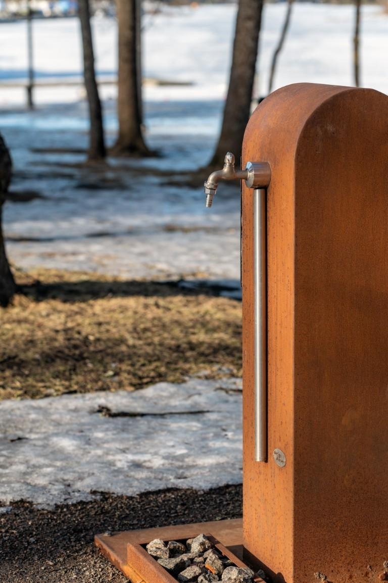 The image shows an outdoor water tap attached to a rustic, metal post. The ground around it has some stones and is partially covered with snow. The background features a few trees and a snowy landscape.