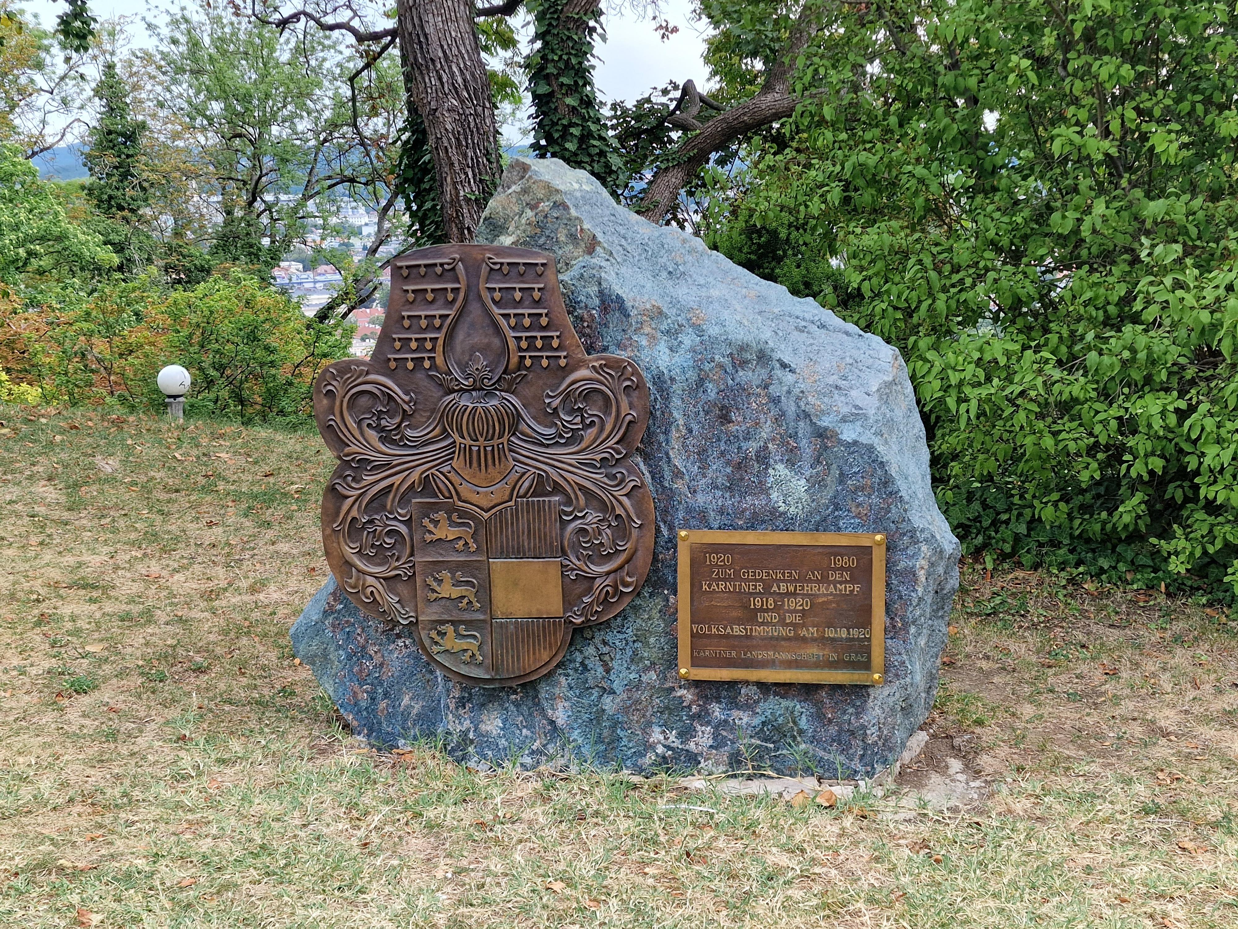 A quiet piece of history with this Gedenkstein (memorial stone) tucked away in the greenery of the Schlossberg. The rough-hewn, blue-grey rock features a large, ornate bronze coat of arms belonging to the Austrian state of Carinthia (Kärnten), easily recognized by its three lions. Next to it, a gold plaque explains its purpose: it commemorates the Kärntner Abwehrkampf (Carinthian Defensive Struggle) of 1918–1920 and the historic plebiscite on October 10, 1920, when the region voted to remain part of Austria rather than joining the Kingdom of Serbs, Croats, and Slovenes. Erected in 1980 by the Carinthian community in Graz, the monument sits peacefully on the grass, framed by mature trees with a glimpse of the city in the background, offering a solemn reminder of post-WWI border conflicts amidst the park's calm.