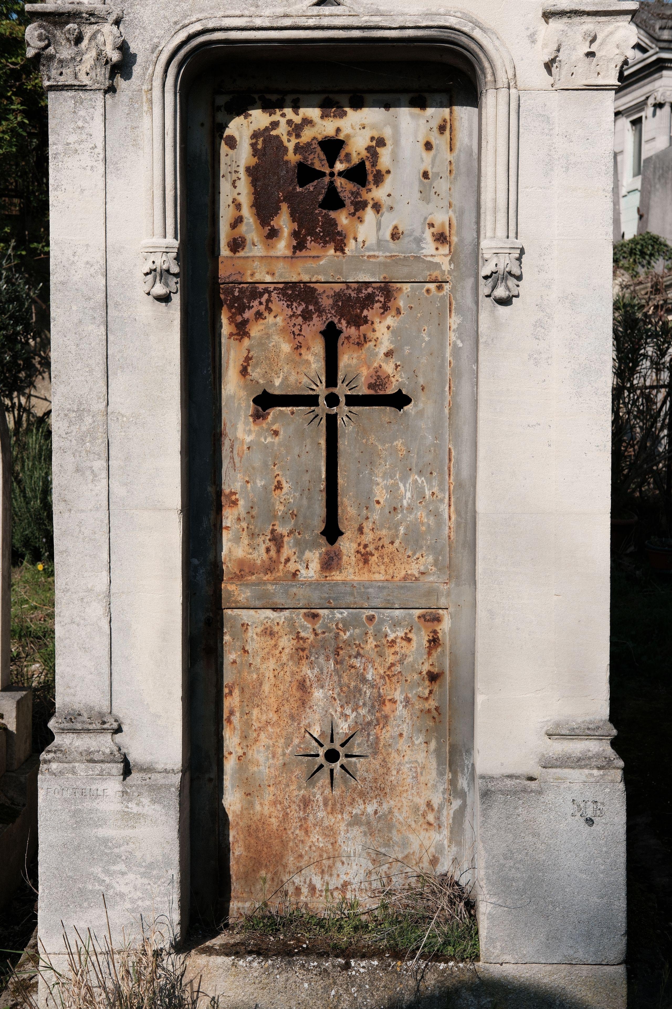 Photo verticale. Façade d’un mausolée avec une porte en métal gris, avec des croix coupées dans le métal. De la rouilles et présente un peu partout