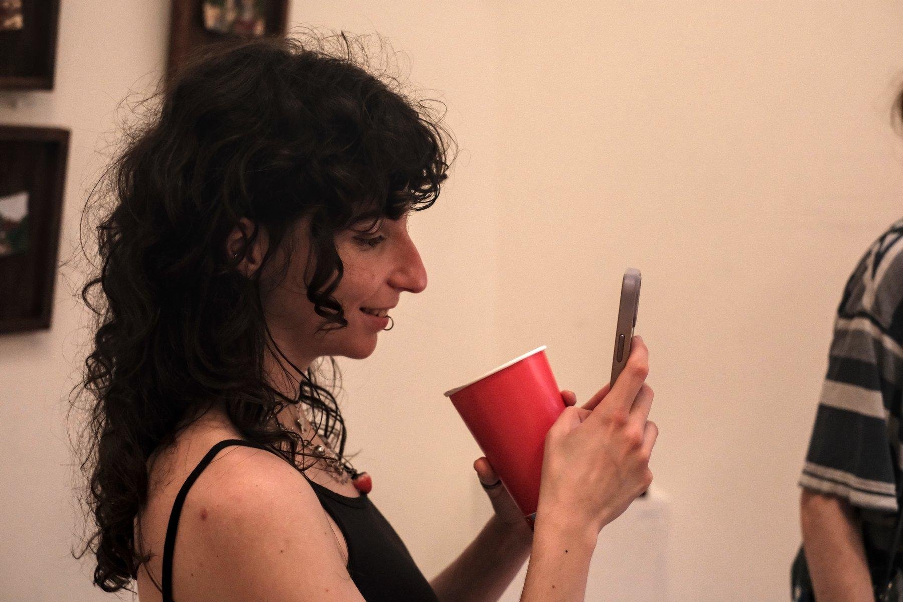 Photo horizontale. Une jeune femme au cheveux brun long et ondulés, de profil, un gobelet rouge à la main gauche et tenant son téléphone de la main droite, en train de filmer la scène devant elle avec un sourire de satisfaction