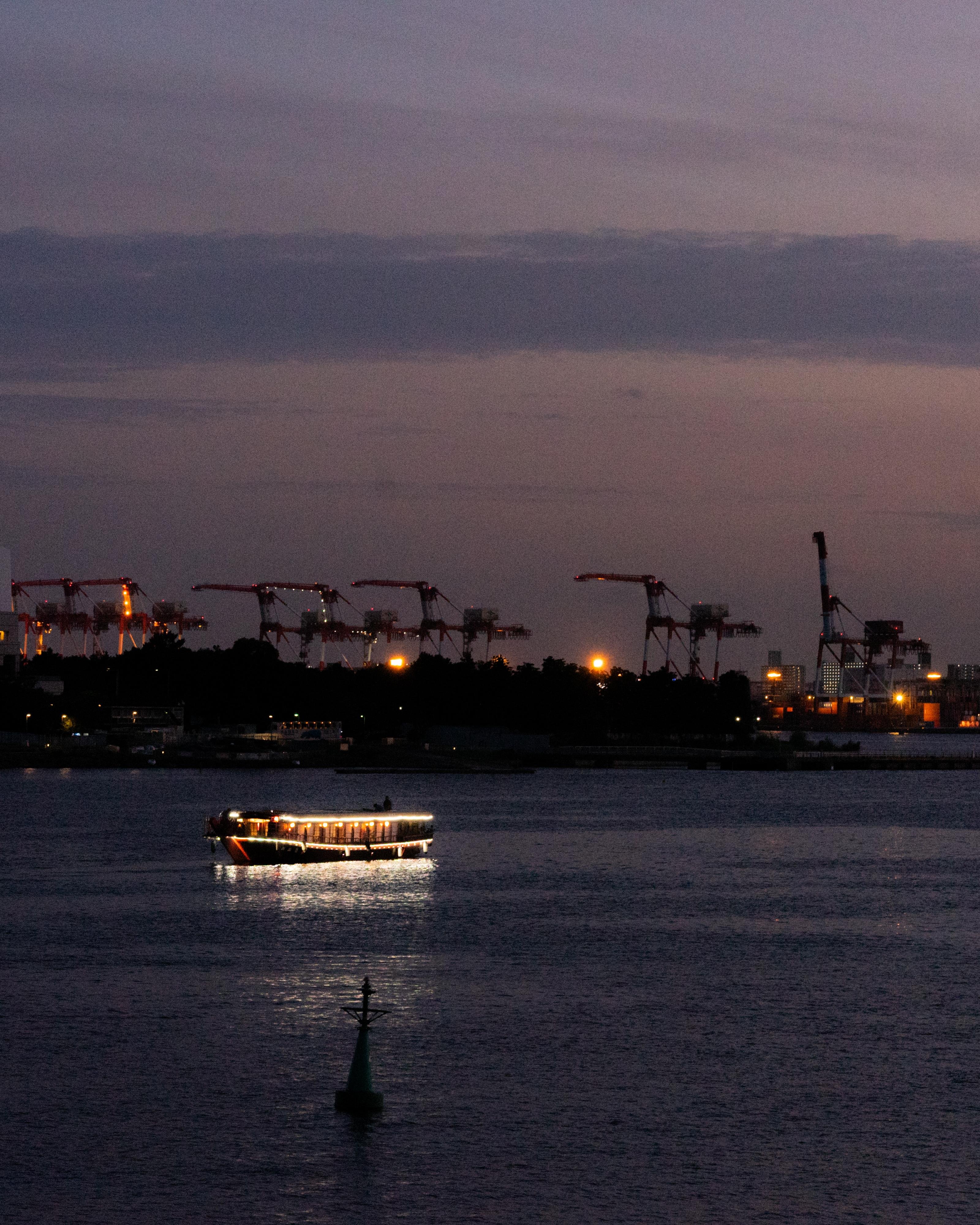 Boat illuminated with lights on the water at dusk, with industrial cranes and city lights visible in the background against a dusky sky. A buoy is present in the foreground.