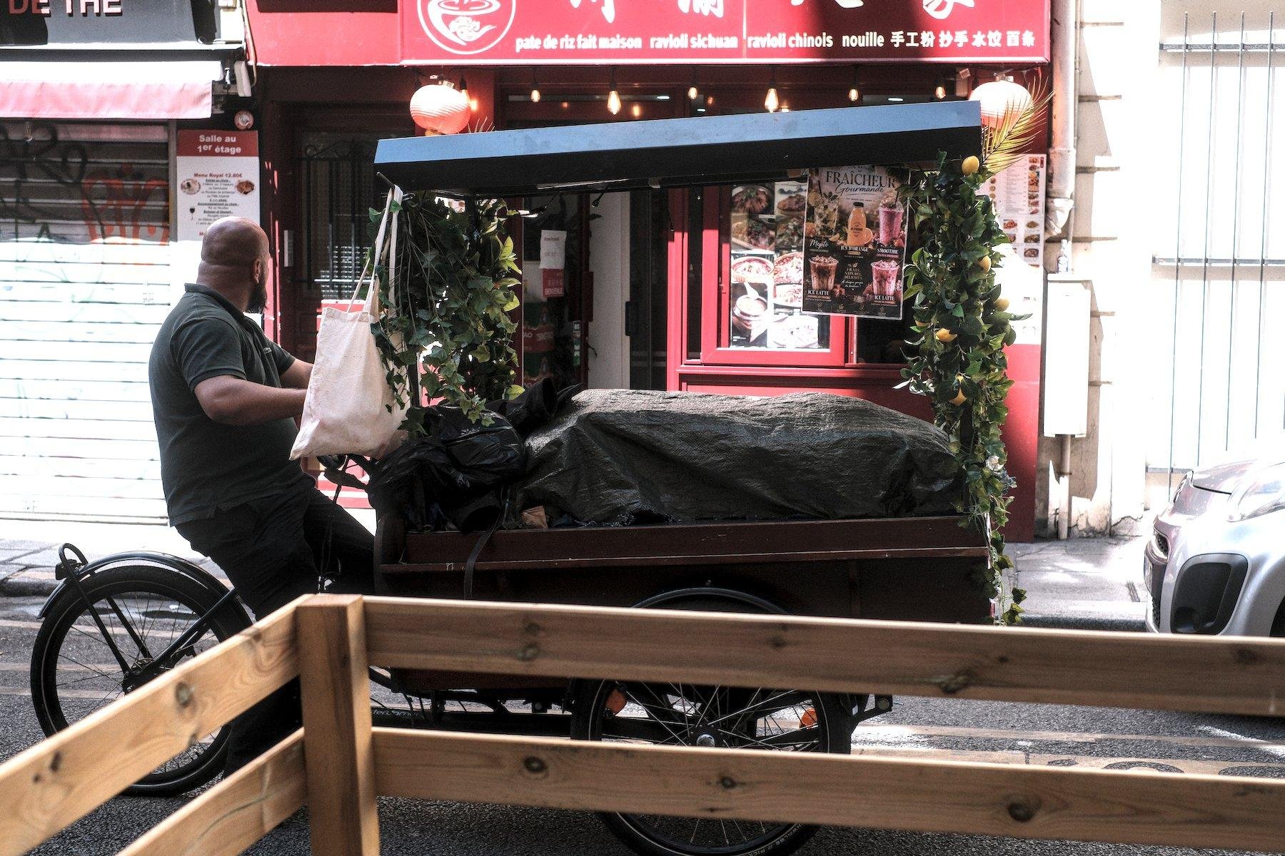 Photo horizontale. Un homme chauve et barbu, bien baraqué, sur un vélo avec un grand cargo à l’avant, protégé par un petit toit bleu. Le contenu est sous une bâche. Il tourne la tête vers sa gauche, à l’opposé de la caméra, vers un restaurant chinois à la façade rouge éclairée par le soleil