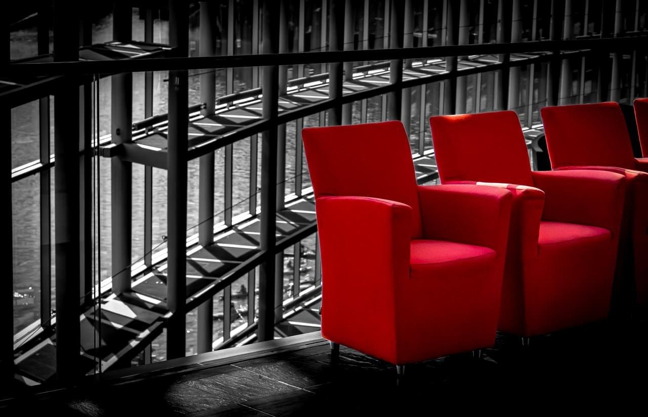 Red chairs aligned in a row, positioned near a large glass window with a view of an urban setting. The background features a monochrome depiction of building structures and water, creating a stark contrast with the vibrant red upholstery of the chairs.