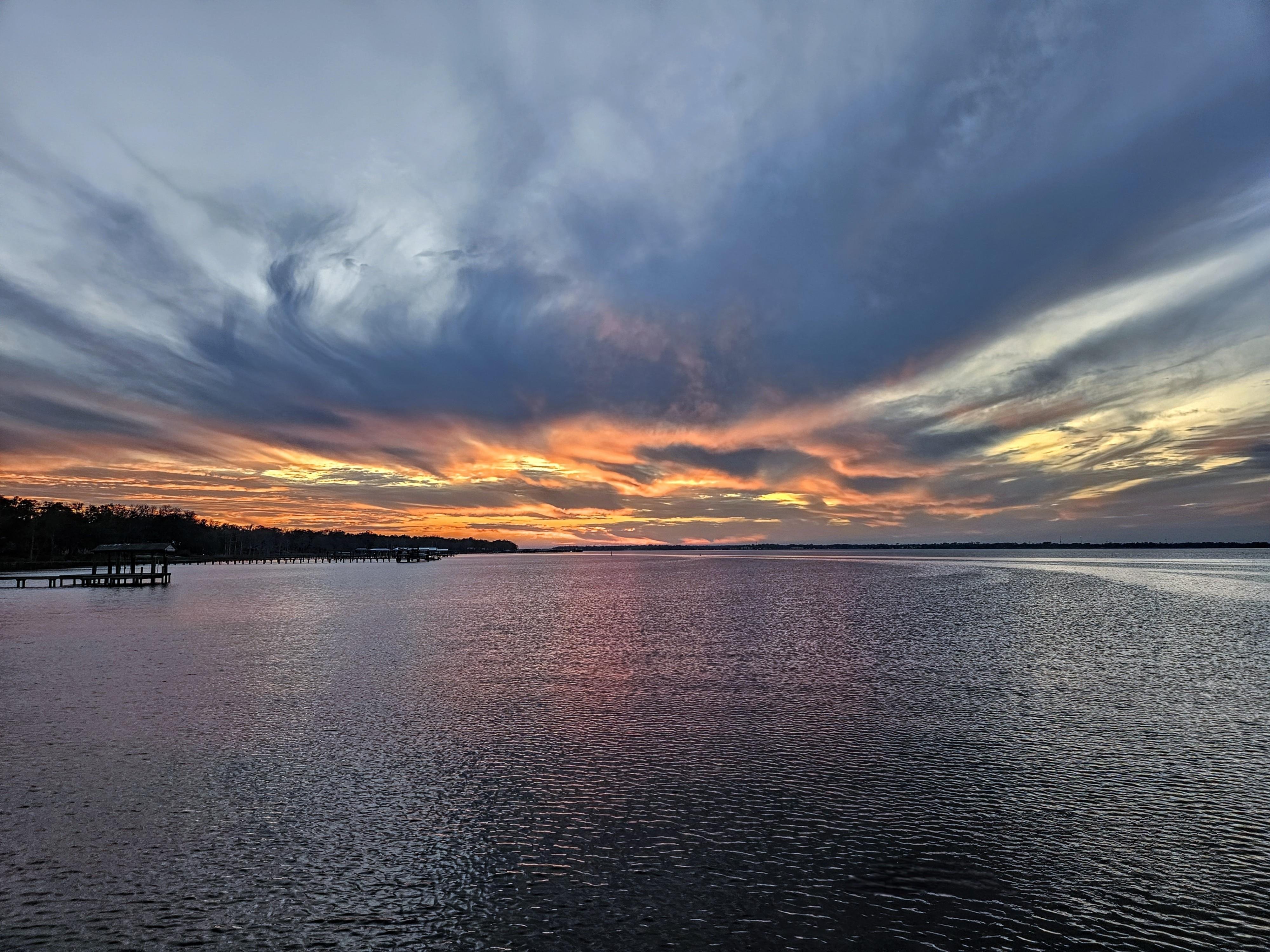 Dramatic sunset over a large body of water, with vivid orange and pink hues illuminating the sky amid dark, swirling clouds. A silhouette of a pier extends from the left edge, while a distant tree-lined horizon is visible. Rippling water reflects the colorful sky.