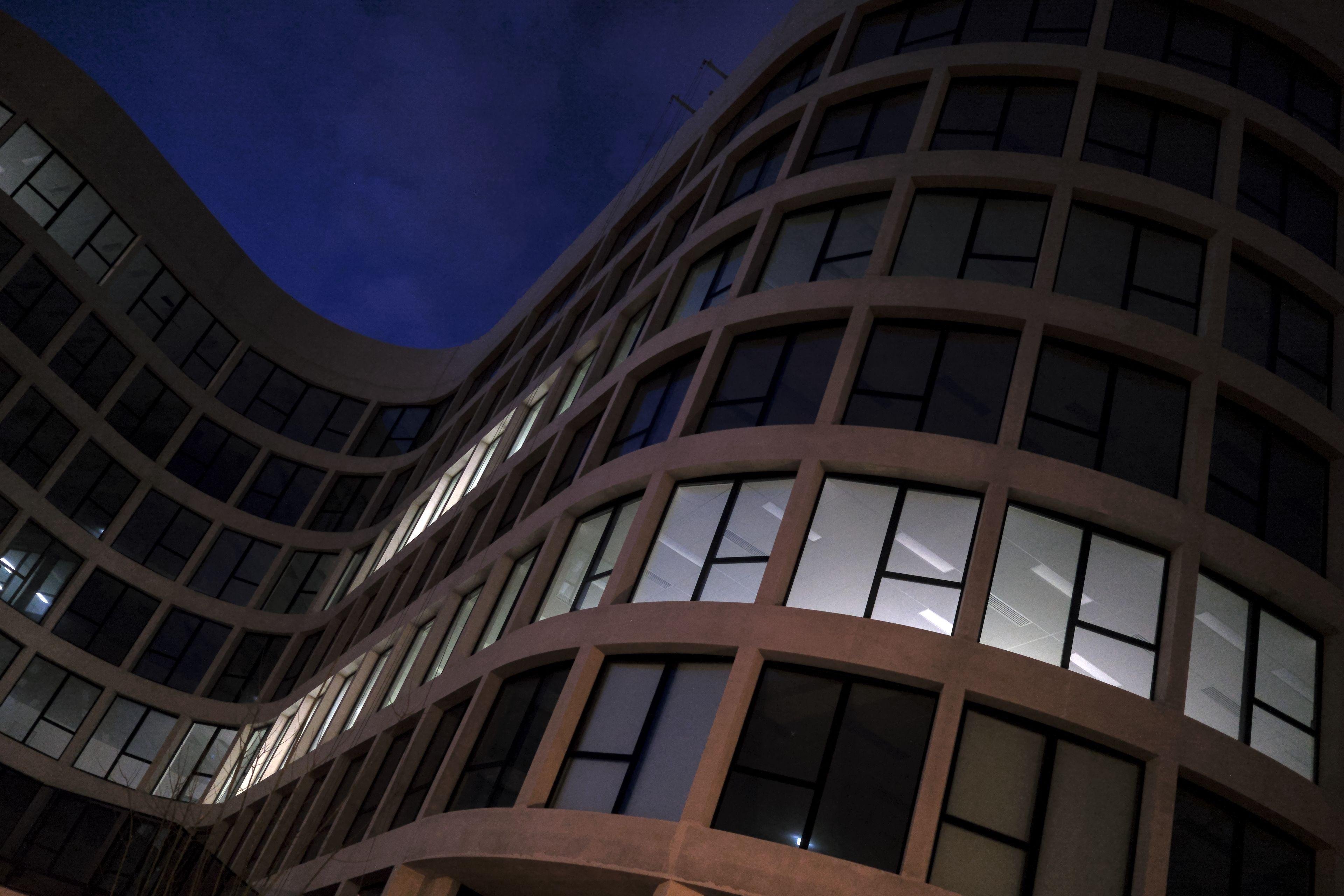 Photo horizontale de nuit. Contre-plongée sur un bout d’immeuble moderne tout en courbes, un bout de ciel bleu nuit apparaît dans le creux de la courbe dessinée