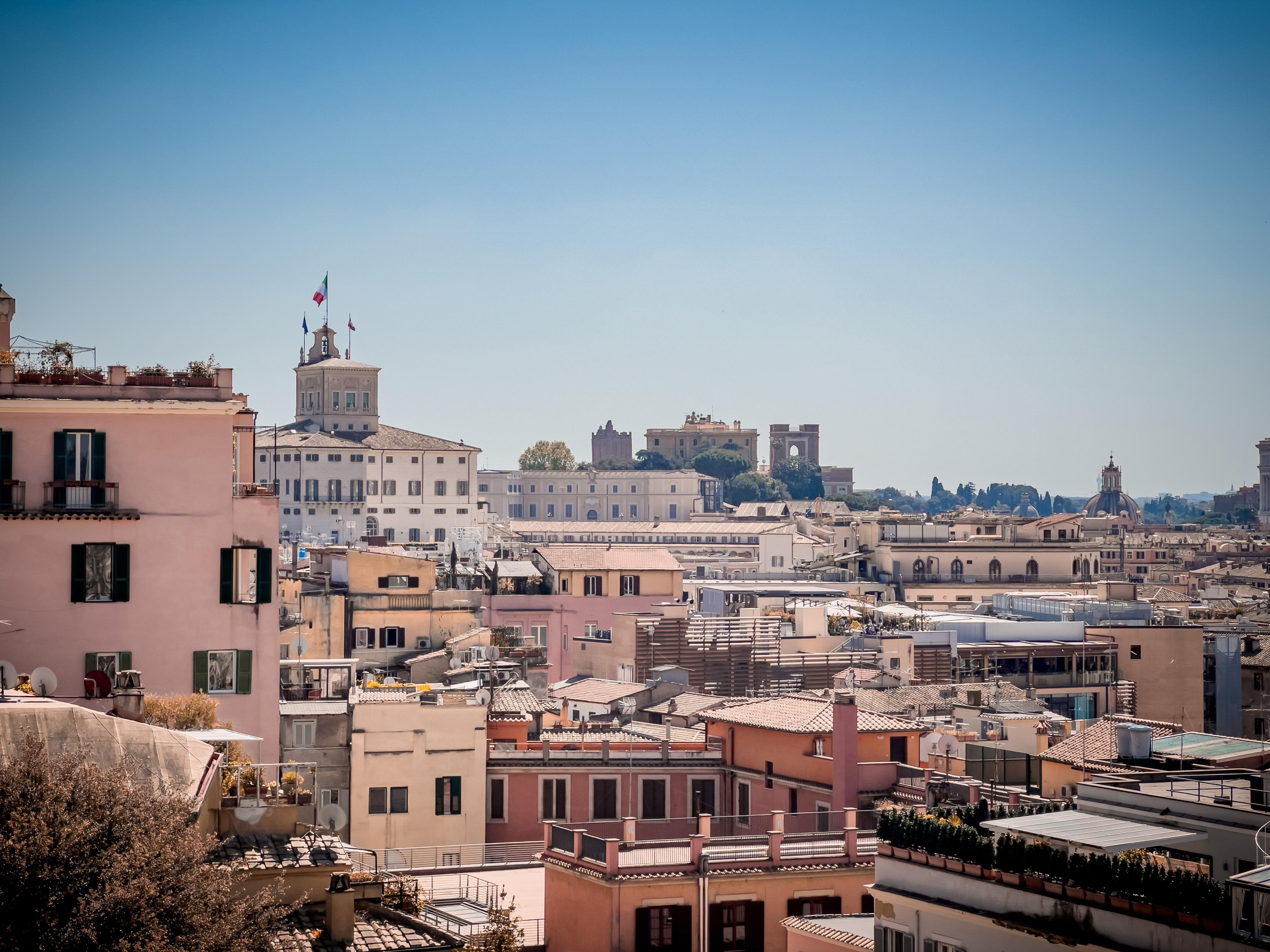 Paesaggio urbano con un mix di edifici storici e moderni. Ci sono diversi edifici a più piani con tetti piatti e, sullo sfondo, si può vedere un edificio prominente con delle bandiere in cima. L'ambientazione sembra quella di una vecchia città europea, probabilmente con punti di riferimento che suggeriscono un'importanza storica. Il cielo è limpido e azzurro, indicando una giornata soleggiata.