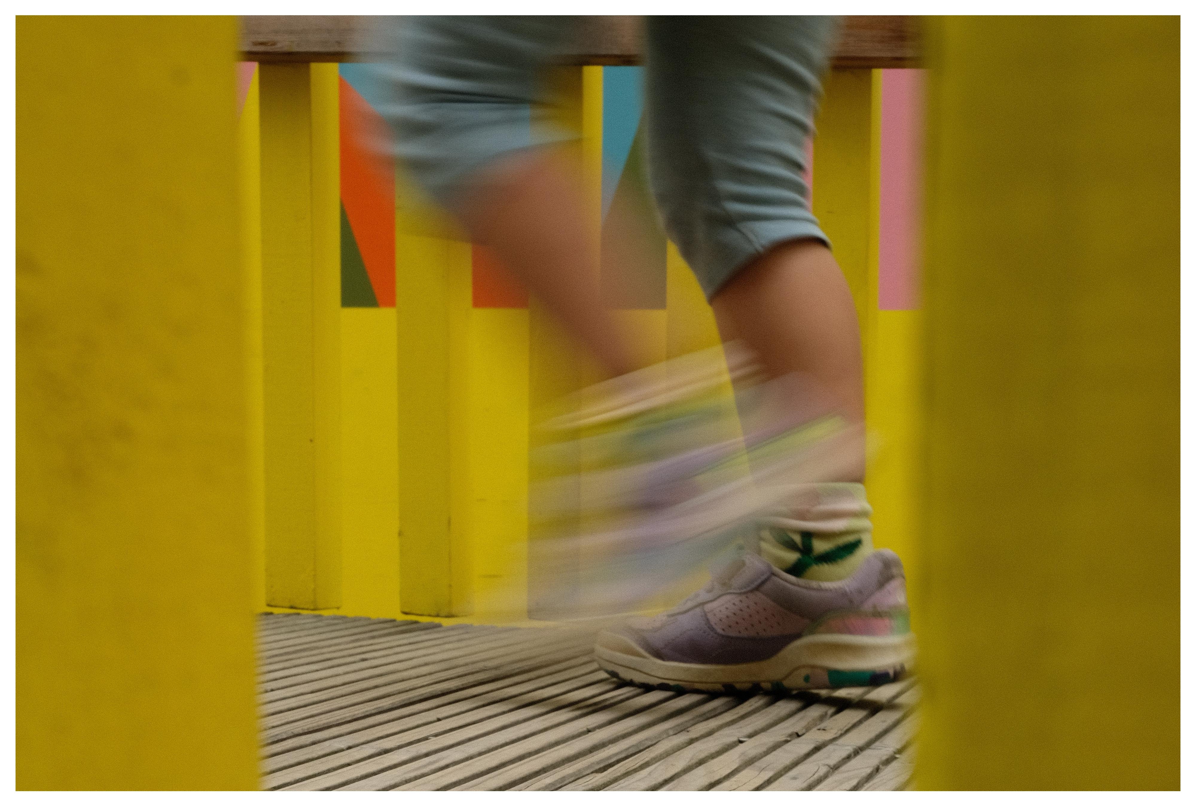Blurred legs of a child in cropped jeans and sneakers running across a slatted wooden deck, framed by bright yellow vertical posts with colorful panels in the background.