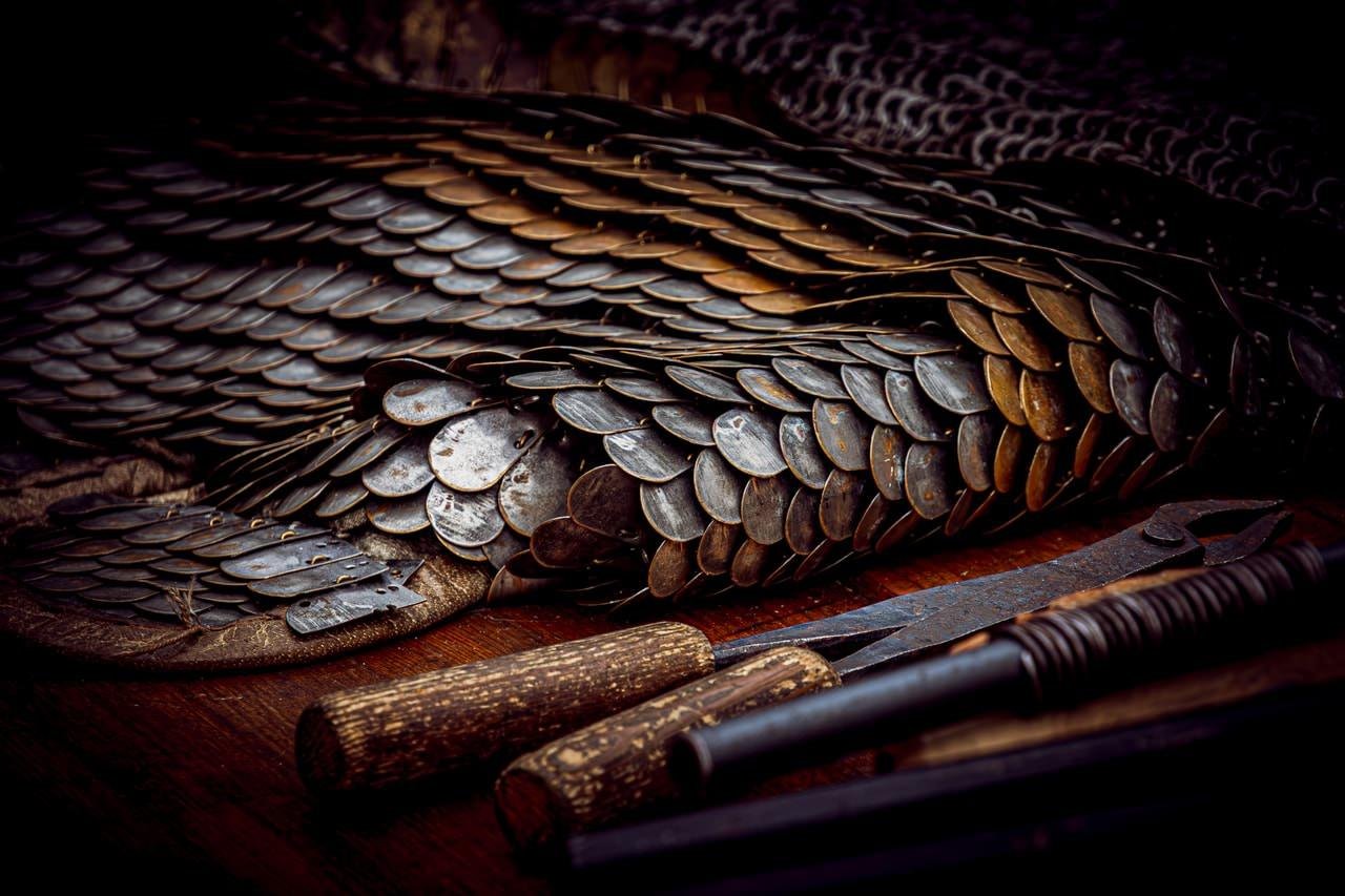 Close-up of overlapping metal scale armor on a wooden surface, with a knife and two cylindrical grips in the foreground under low, warm lighting.