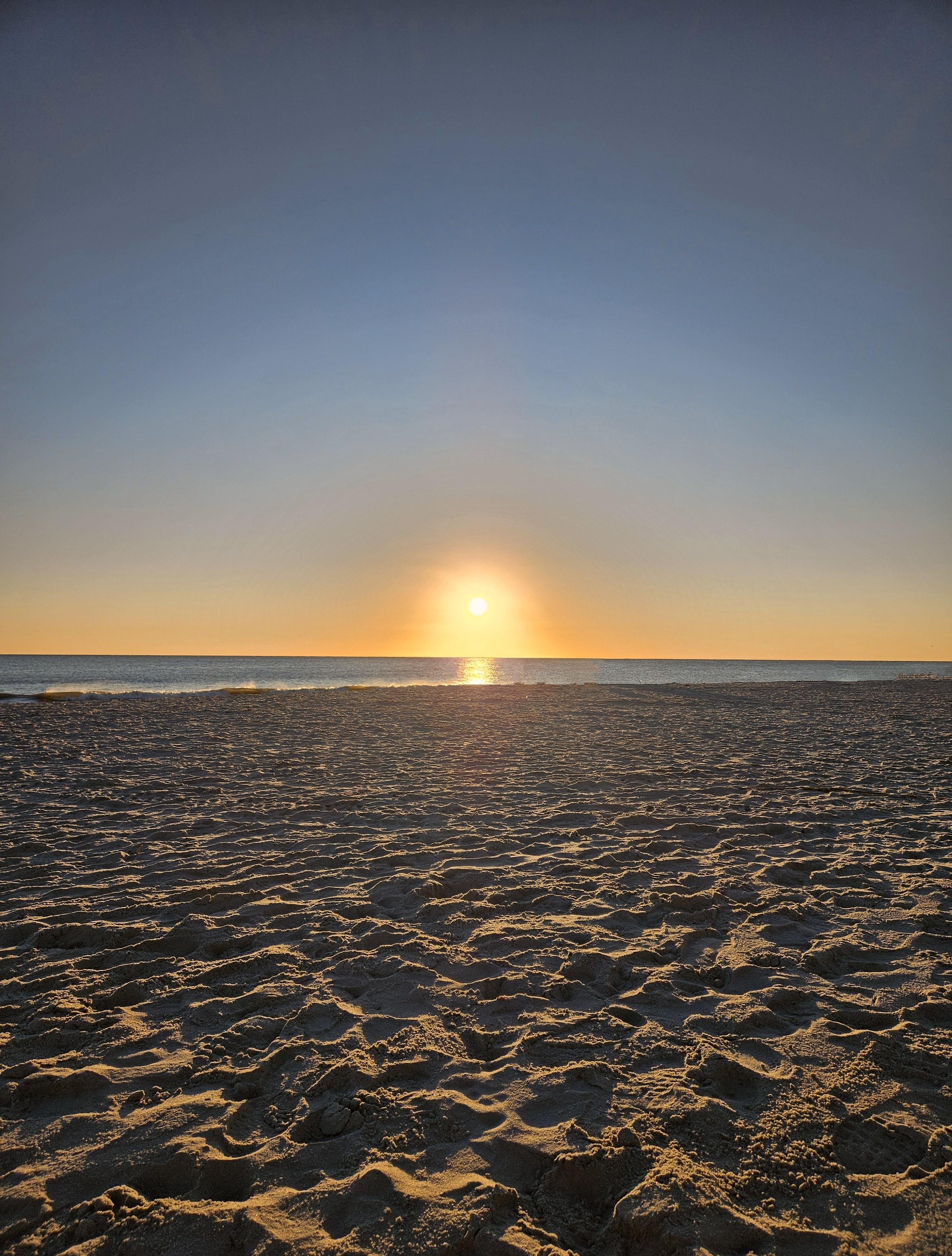 Sunrise over a beach with the sun low on the horizon, casting a warm glow on the ocean and sky. The foreground features textured sand with shadows highlighting ripples and footprints. The horizon line is straight and uninterrupted, dividing the sky and sea.
