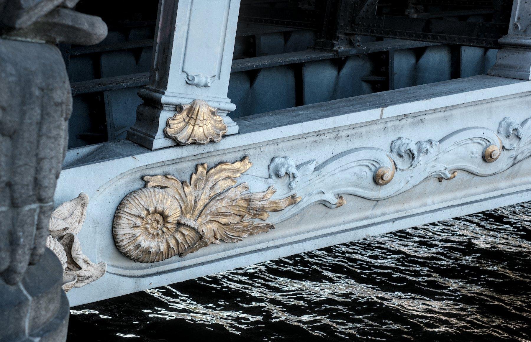 Photo horizontale. Gros plan sur un détail de la structure d’un pont, gris clair, avec des motifs sculptés et certains couvert de peinture dorée :  une coquille de créature marine