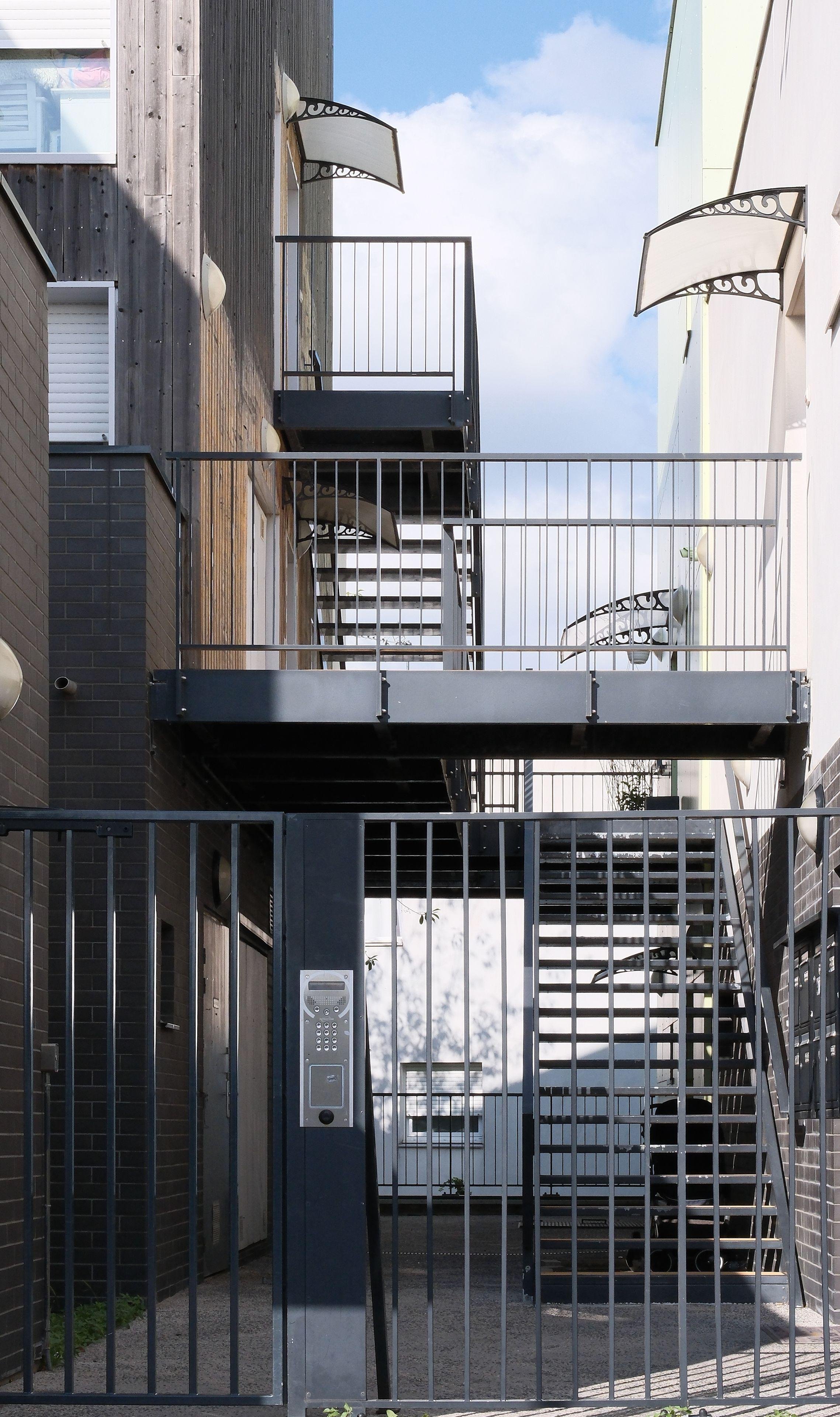 Photo verticale. Des escaliers desservant 2 petits immeuble de 3 étages à gauche et à droite, derrière un portail à digicode. L’immeuble de gauche est en finition bois et celui de droite en peinture blanche