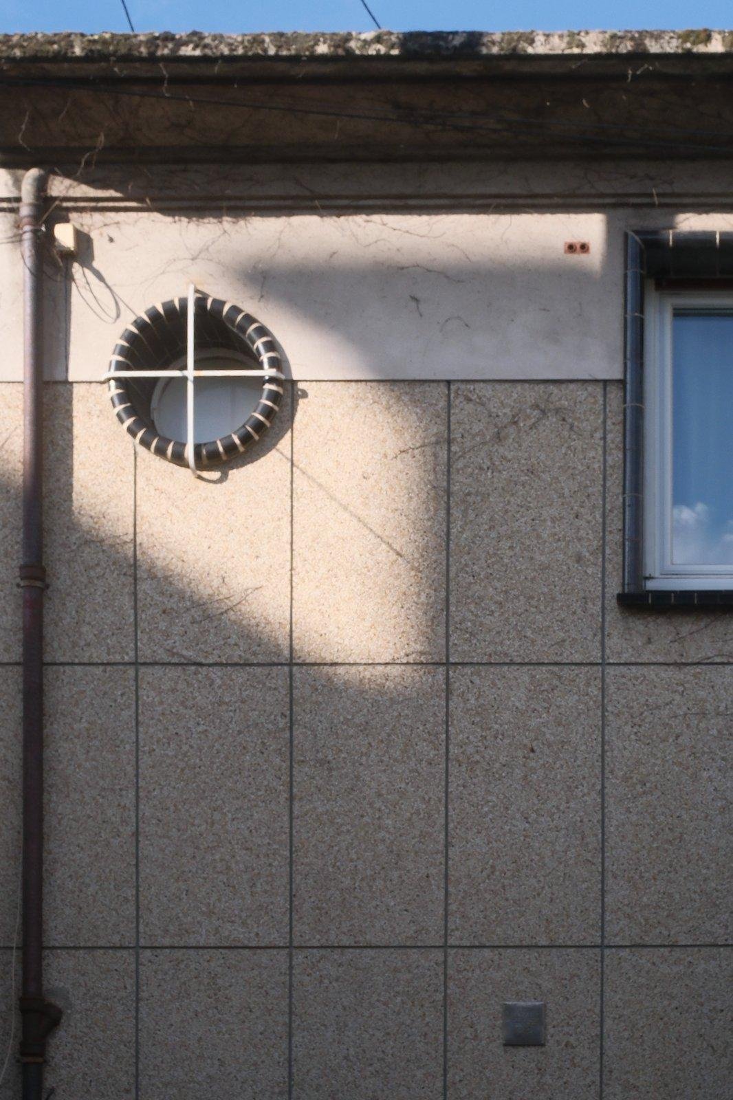 Photo format portrait, partie de la façade d’une maison, avec une fenêtre toute ronde cerclée avec une décoration noire avec des traits blanc, évoquant une bouée, et barrée d’une croix en métal formant le signe plus