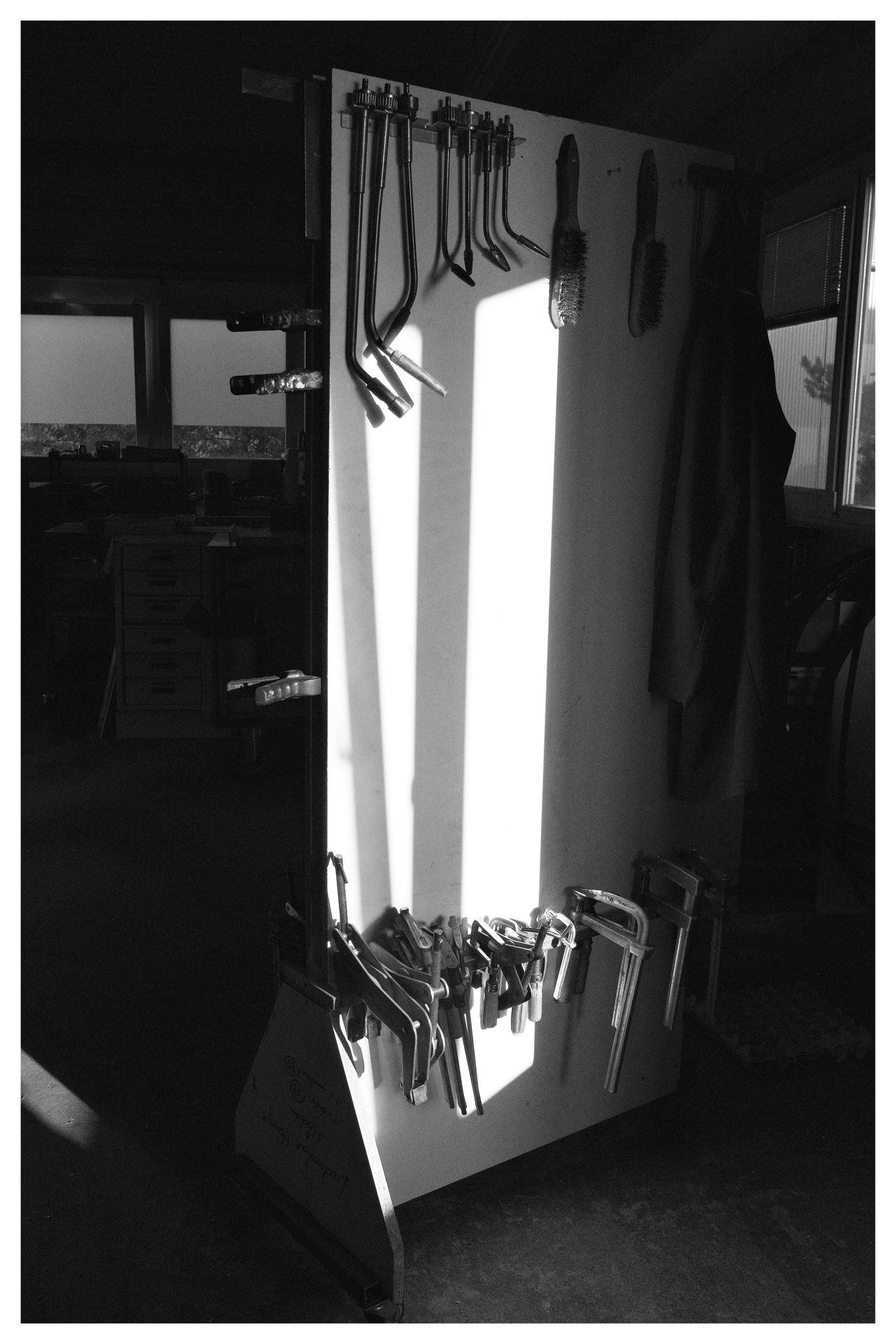 A monochrome image of a tool storage area with assorted metal clamps hanging on a vertical board. Several long-handled tools and brushes are mounted above the clamps. Sunlight casts strong shadows on the board and the floor. To the left, a window with partially closed blinds is visible in the background, and to the right, a heavy fabric hangs on the side.