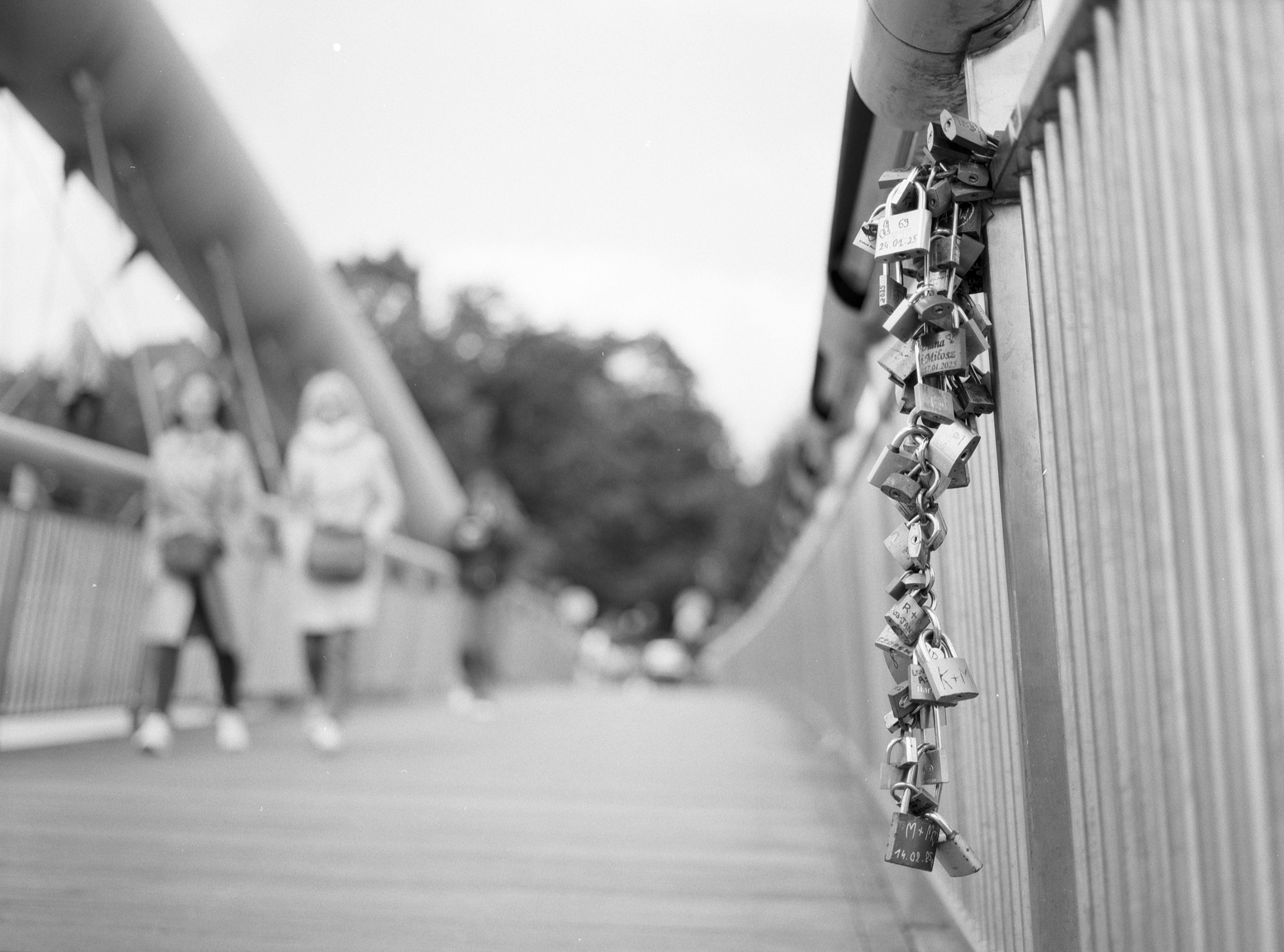 Black and white photograph of a bridge with numerous padlocks attached to the railing on the right side. Two blurred figures walk on the bridge in the background. Vertical poles line the bridge on both sides, converging towards a distant, out-of-focus area. Trees and an overcast sky appear in the background.