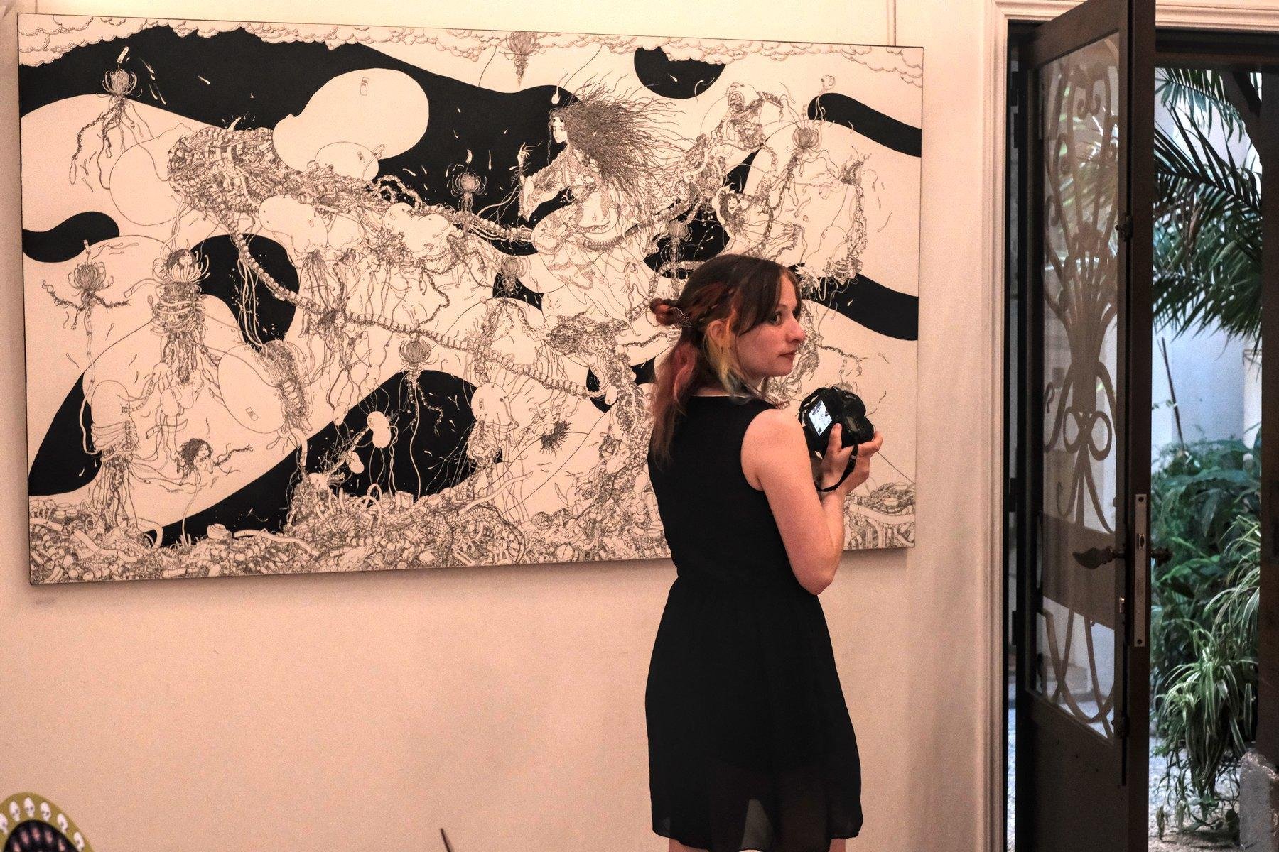 Photo horizontale. Devant une grande toile avec une œuvre en noir et blanc très détaillée, une jeune femme en robe noire, cheveux colorés et coiffés, se tourne sur la droite pour regarder derrière elle, tenant un appareil photo dont l’écran est allumé