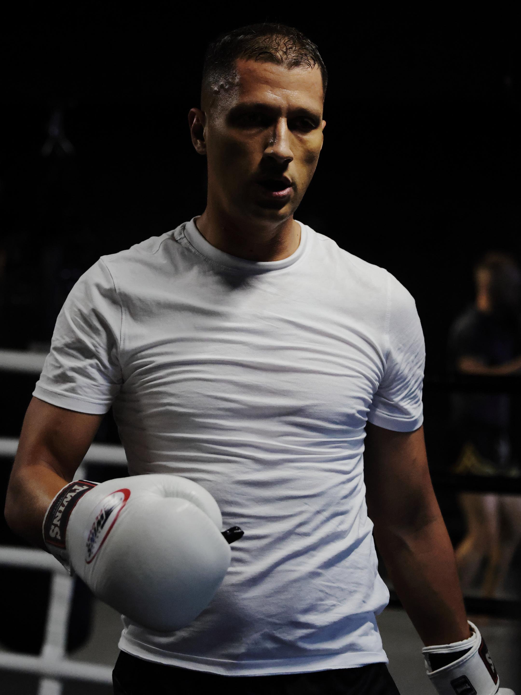 Male boxer wearing a white t-shirt and white boxing gloves, standing in a dimly lit boxing ring. His face and shirt are slightly sweaty, indicating recent physical activity. The background is dark with indistinct shapes of people or objects.