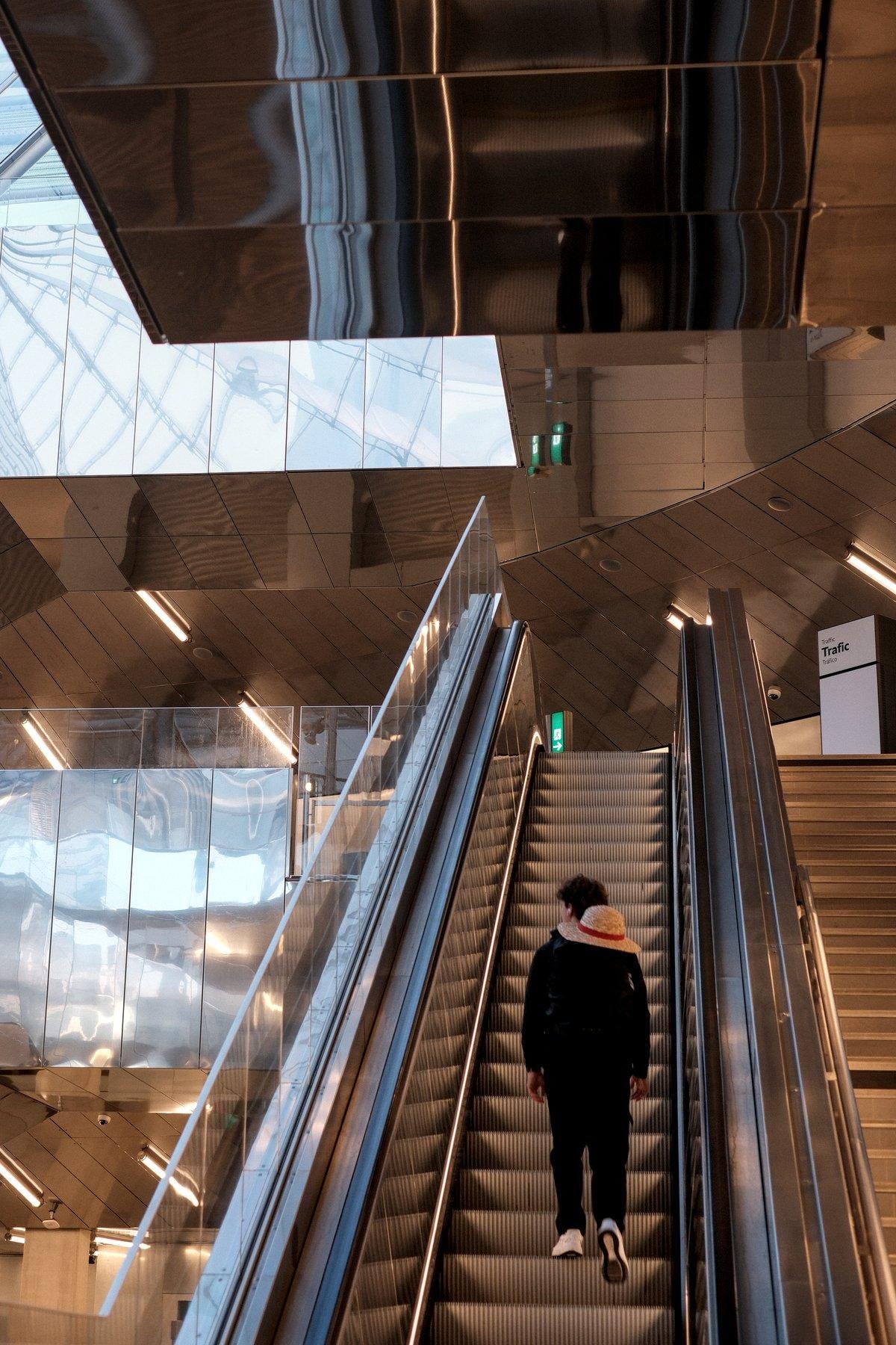 Photo verticale. Sur un escalator, Un homme de dos est en train de le gravir. Il est habillƩ de noir, baskets blanches, et un chapeau de paille avec un bandeau rouge dans le dos