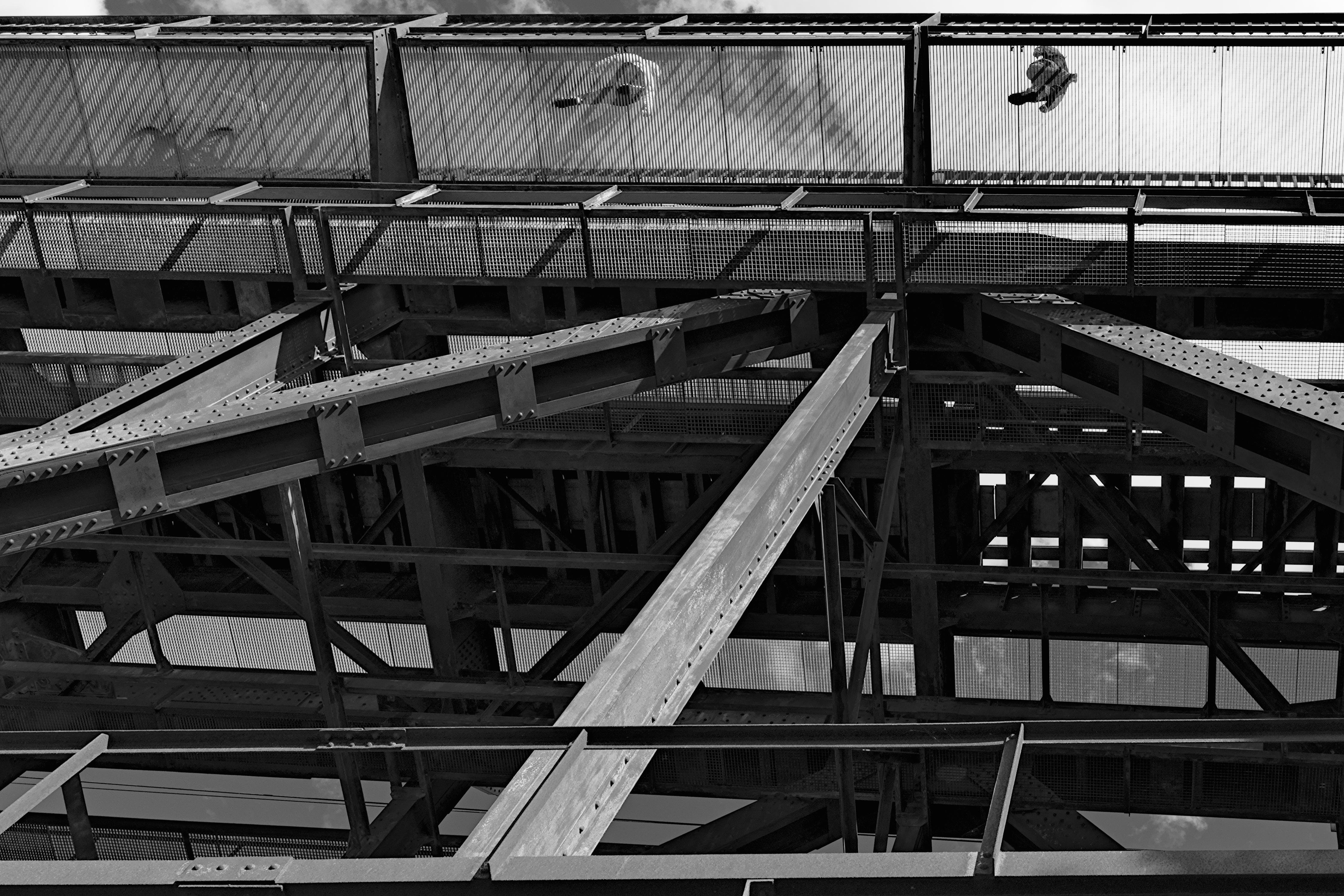 Metal bridge structure viewed from below, featuring intersecting beams and girders. Overhead, two silhouetted figures walk on a mesh walkway, casting shadows. The sky is visible through the open spaces of the framework. Black and white image highlighting the architectural details.
###
Metallbrückenstruktur von unten gesehen, mit sich kreuzenden Balken und Trägern. Über dem Kopf gehen zwei Silhouettenfiguren auf einem Mesh-Gehweg und werfen Schatten. Der Himmel ist durch die offenen Räume des Gerüst sichtbar. Schwarz-Weiß-Bild, das die architektonischen Details hervorhebt.
