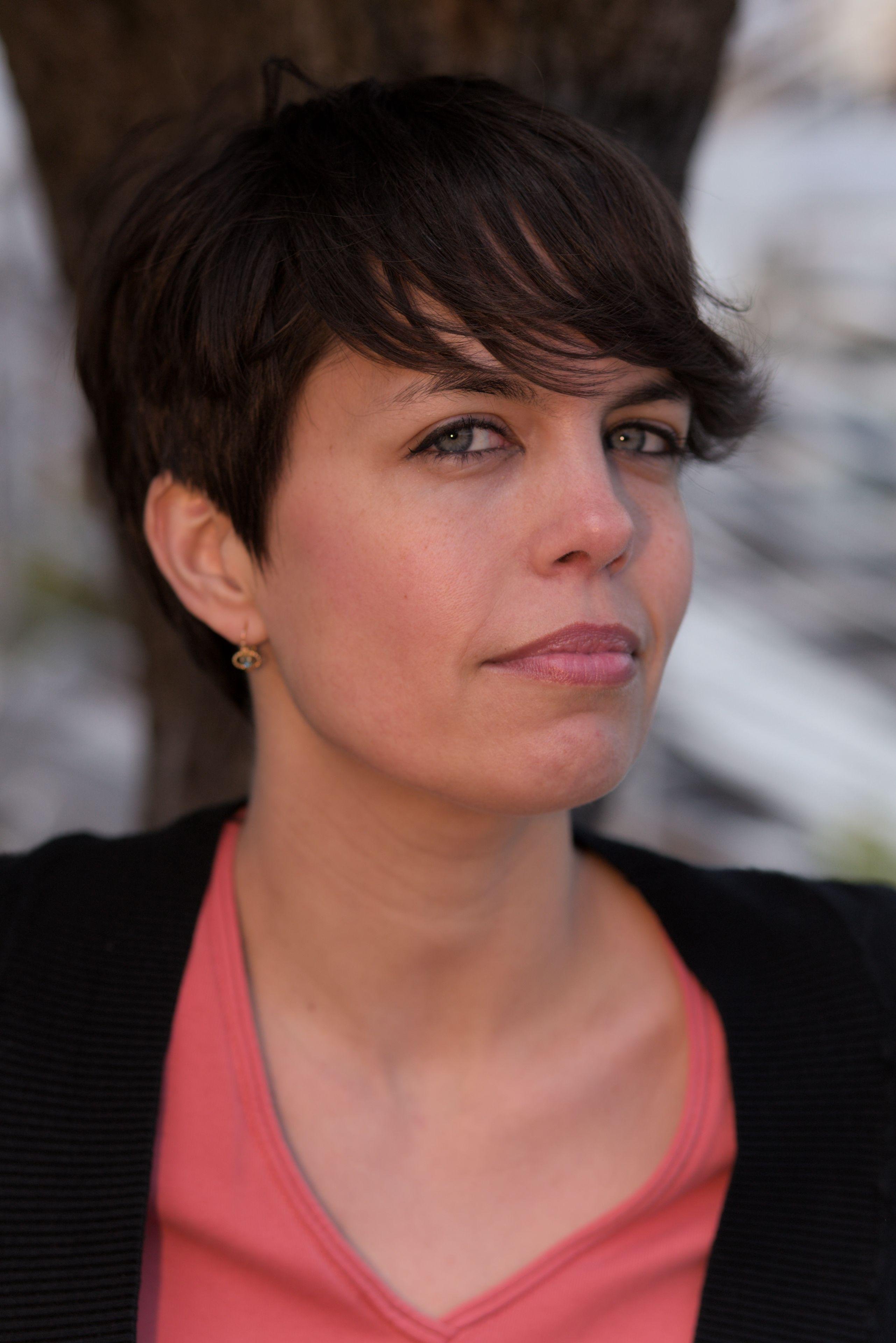 photo verticale. Plan serré sur le visage d’une jeune femme brune qui porte un haut rouge et un gilet noir par dessus. Elle tourne légèrement la tête vers sa gauche et regarde la caméra du coin des yeux