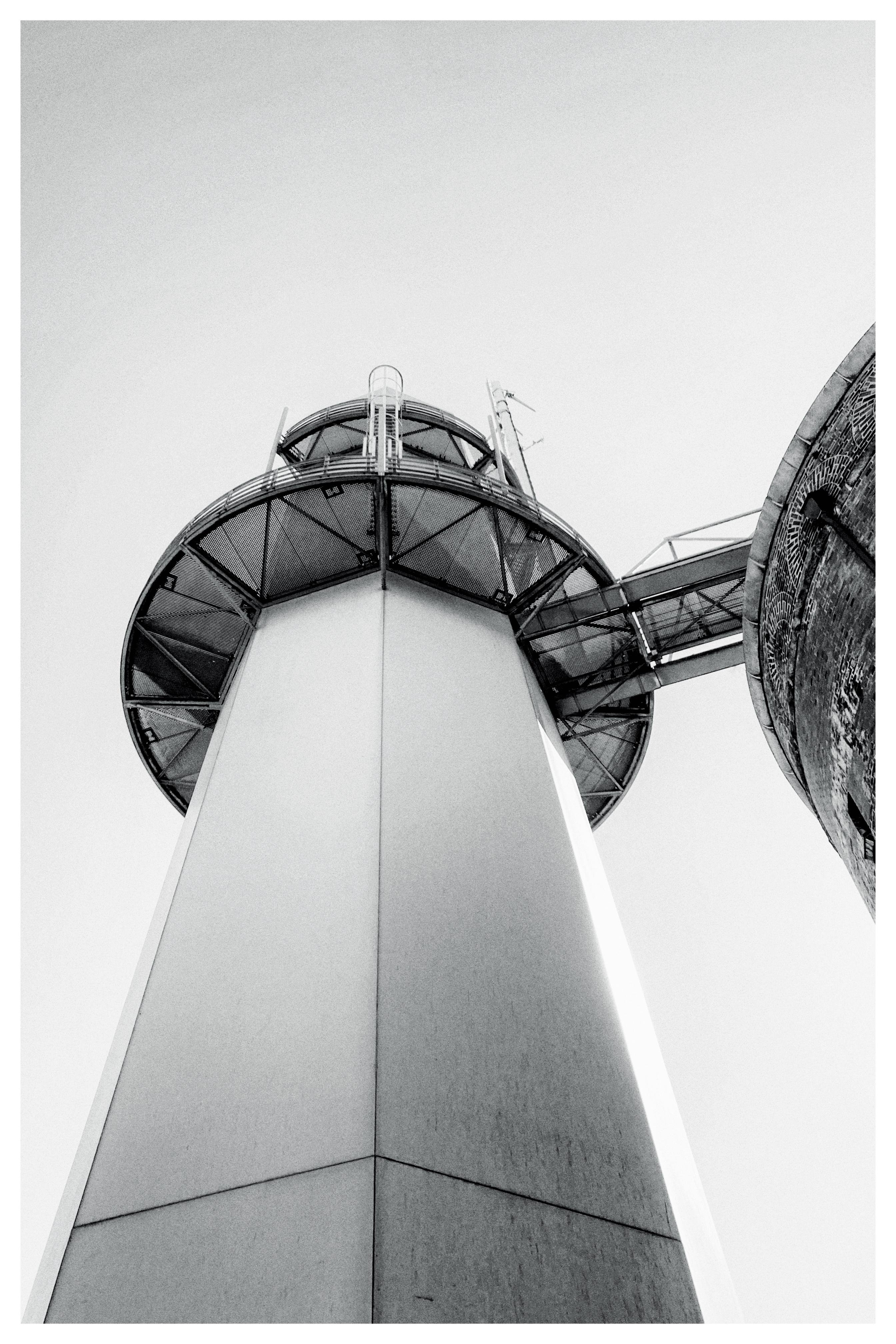 Tall cylindrical structure viewed from below, featuring a wide circular observation deck surrounded by railings at the top. The structure is connected to a second building on the right. Sky appears clear and light in color, suggesting daytime. The perspective emphasizes the height and geometric lines of the tower.