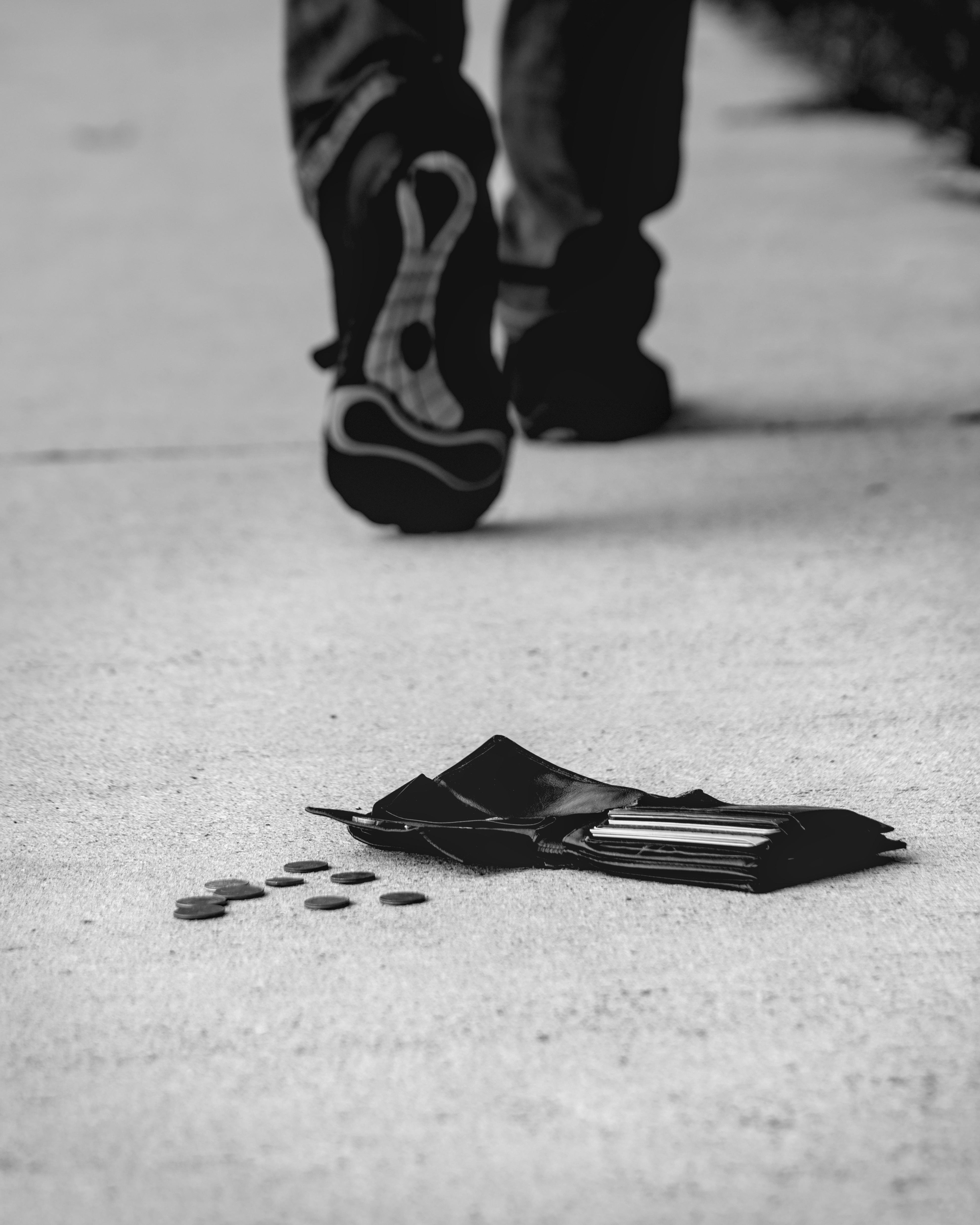 Black-and-white photograph of a sidewalk with a wallet and scattered coins on the ground in the foreground. A person in jeans and athletic shoes is walking away, slightly blurred, in the background.
###
Schwarz-Weiß-Fotografie eines Bürgersteigs mit einer Brieftasche und verstreuten Münzen auf dem Boden im Vordergrund. Eine Person in Jeans und Sportschuhen geht leicht unscharf im Hintergrund weg.