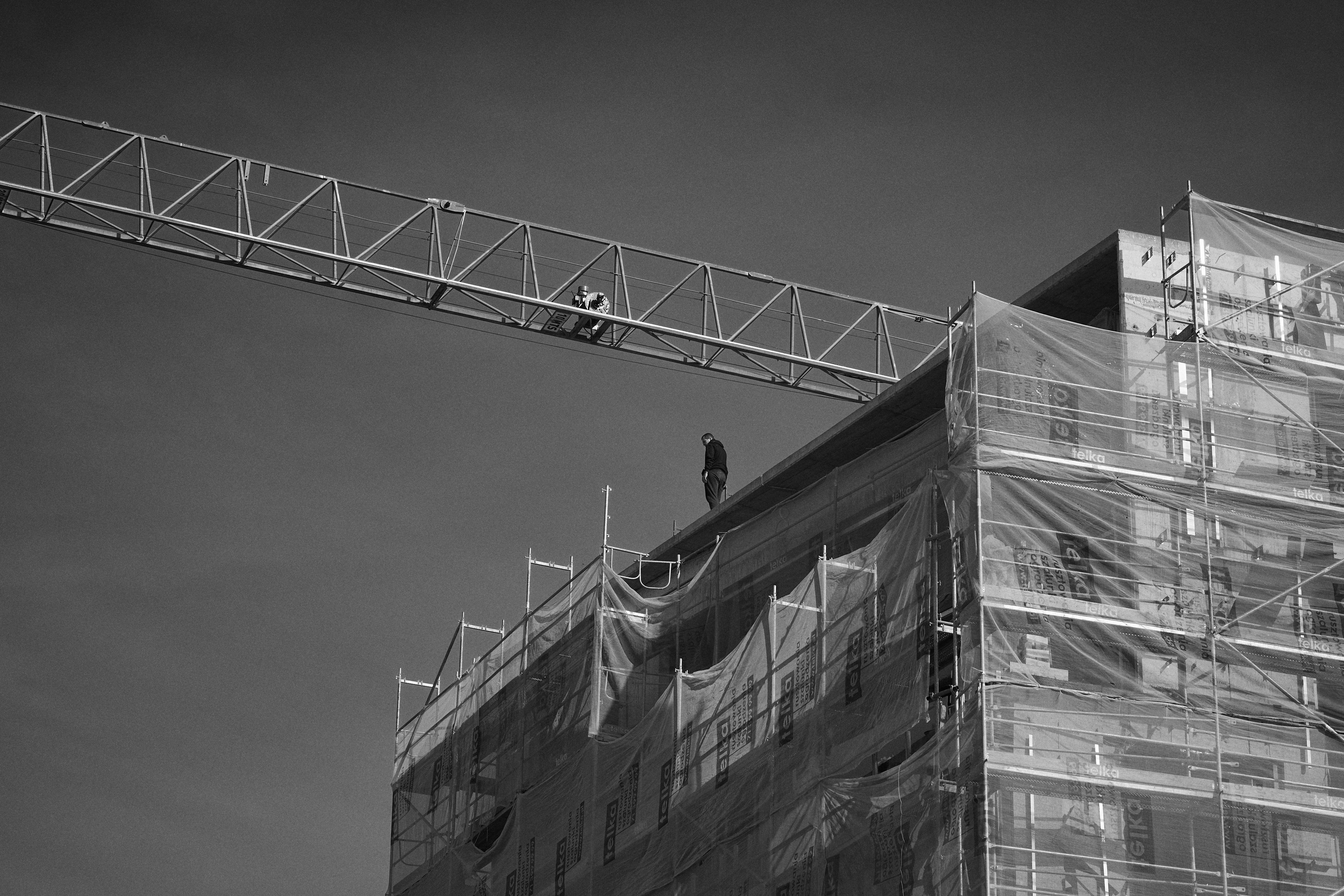 The image shows a construction site with a building covered in scaffolding and protective netting. There is a person standing on the top of the building, and a crane arm is visible above them. The scene is in black and white.