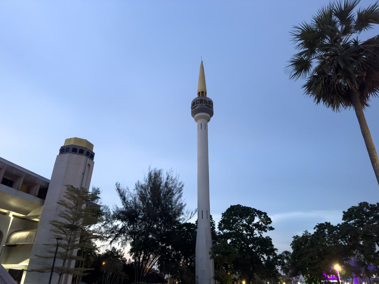 A tall mosque minaret with a gold pointed top stands against a dusk sky. The structure is surrounded by trees, including palm trees on the right. On the left, part of a building with a similar architectural style is visible, with warm lights illuminating its facade.