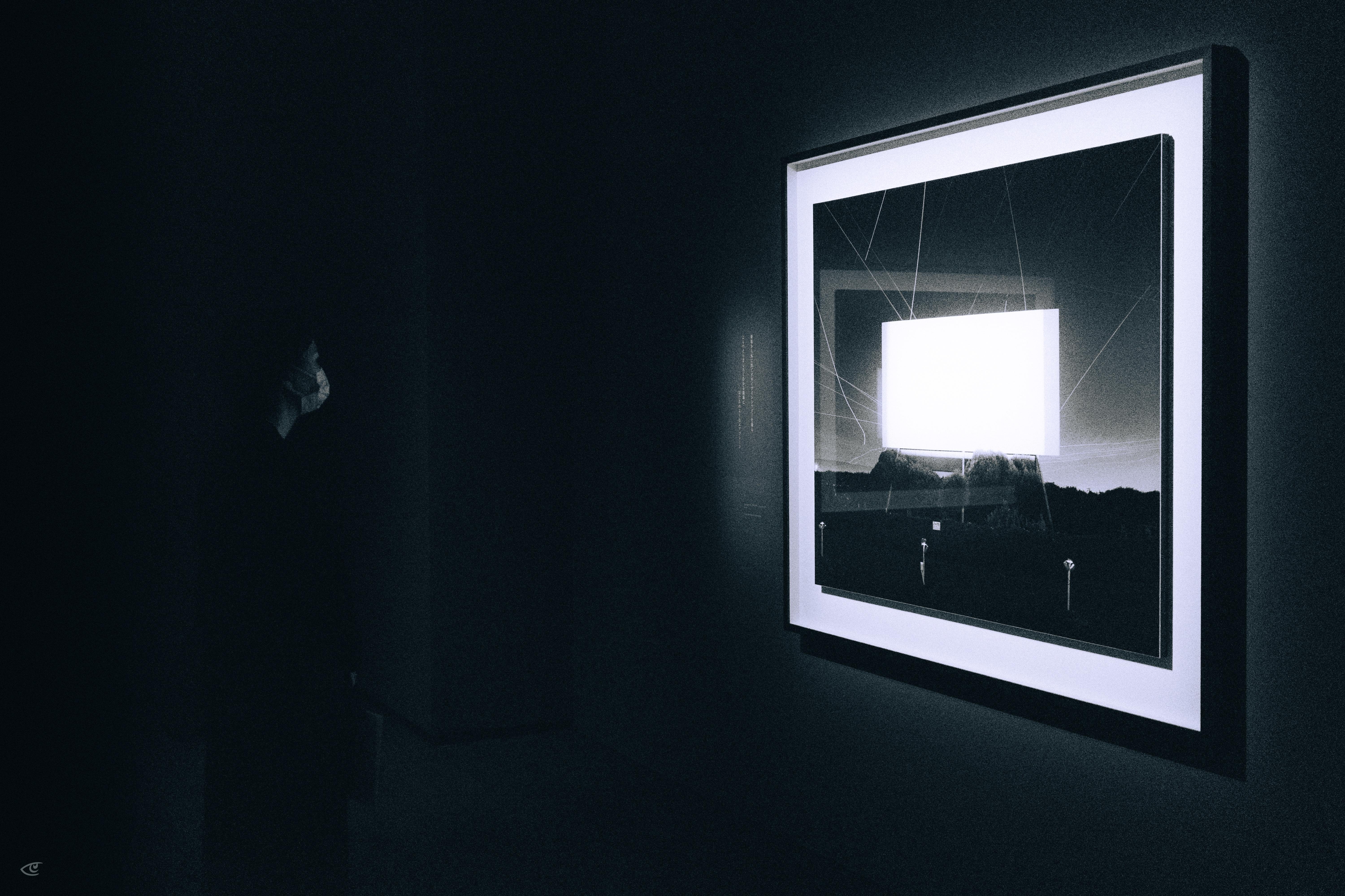Framed photograph on a dark wall, backlit with a glowing white square in the center; faint silhouette of a person at left facing the artwork.