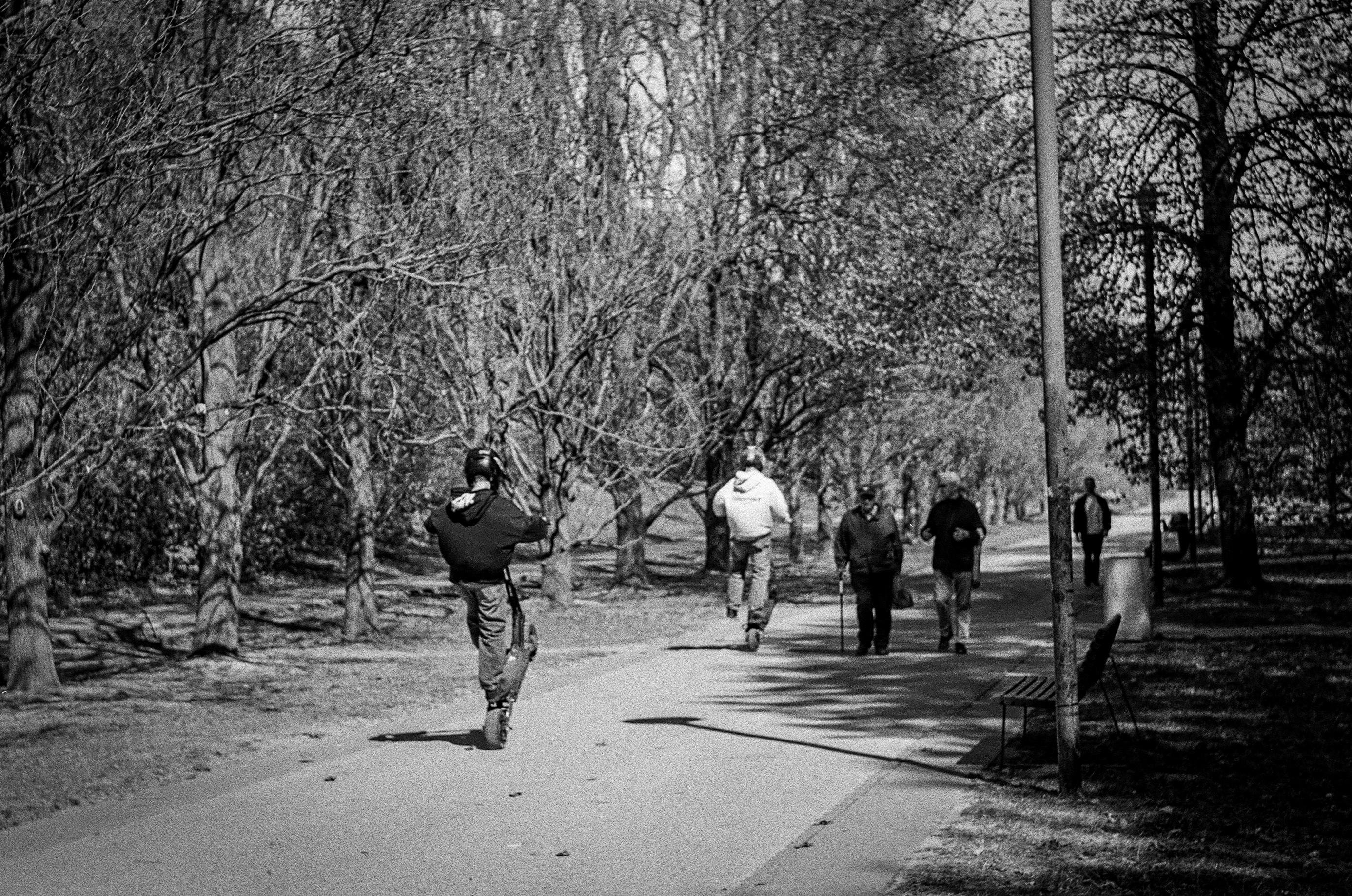 B&W film photo of two kids doing a wheelie on electric scooters in a park. Other people just walk by