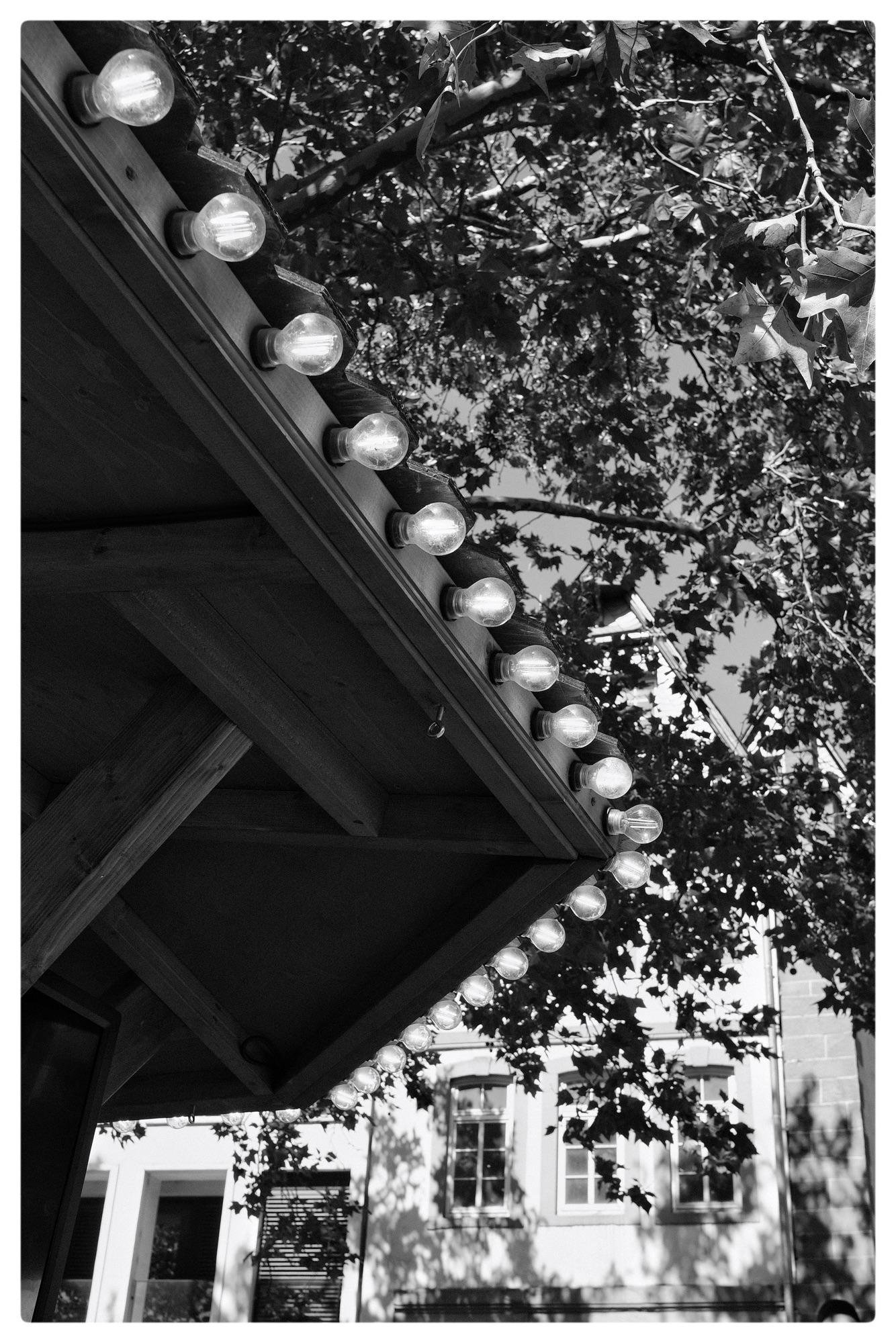 Black and white photo showing a wooden kiosk corner with round light bulbs lining the edge. Tree leaves and branches partially shadow the structure. In the background, a building with rectangular windows is visible through the foliage.