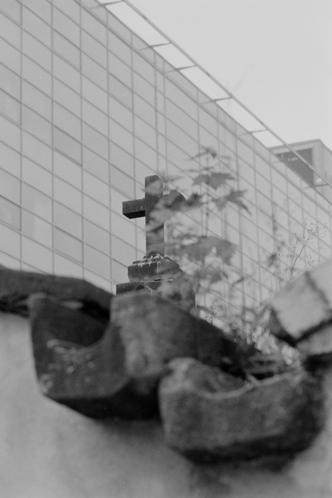 Black-and-white low-angle view of large rocks in the foreground, with a cross-shaped structure partially obscured by foliage and a modern tiled building facade rising behind it.