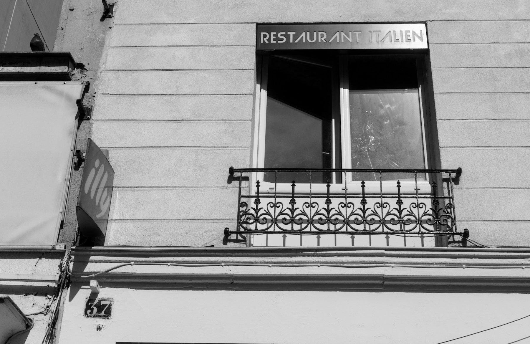 Photo horizontale en noir et blanc. Une fenêtre entre ouverte, avec une enseigne marquée «&nbsp;Restaurant Italien&nbsp;» en haut, sur un mur avec des traits horizontaux dans le béton. Une plaque de rue dans le coin inférieur gauche est marquée «&nbsp;37&nbsp;»