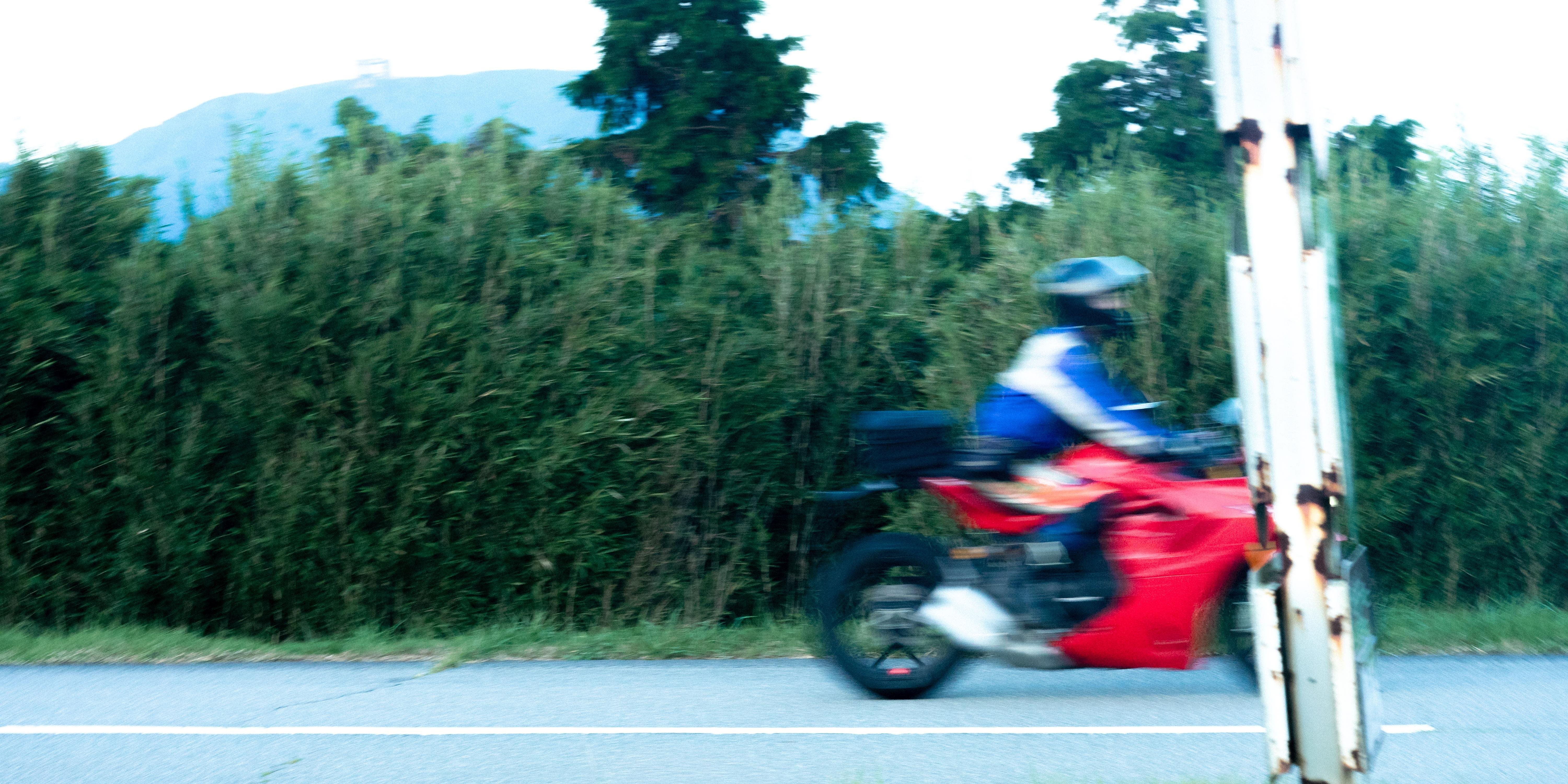 Blurred image of a person riding a red motorcycle past a tall hedge on a road. The rider wears a blue jacket and helmet. A rusty metal pole is visible to the right, with trees and a mountain in the distant background.