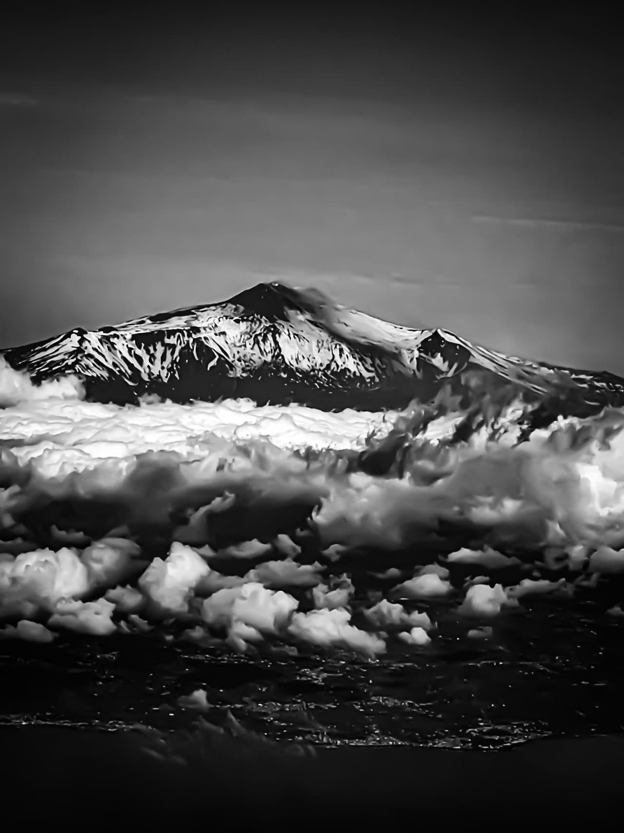 Black-and-white landscape of a snow-covered mountain peak rising above a thick layer of clouds, with darker cloud formations in the foreground and an overcast sky above.