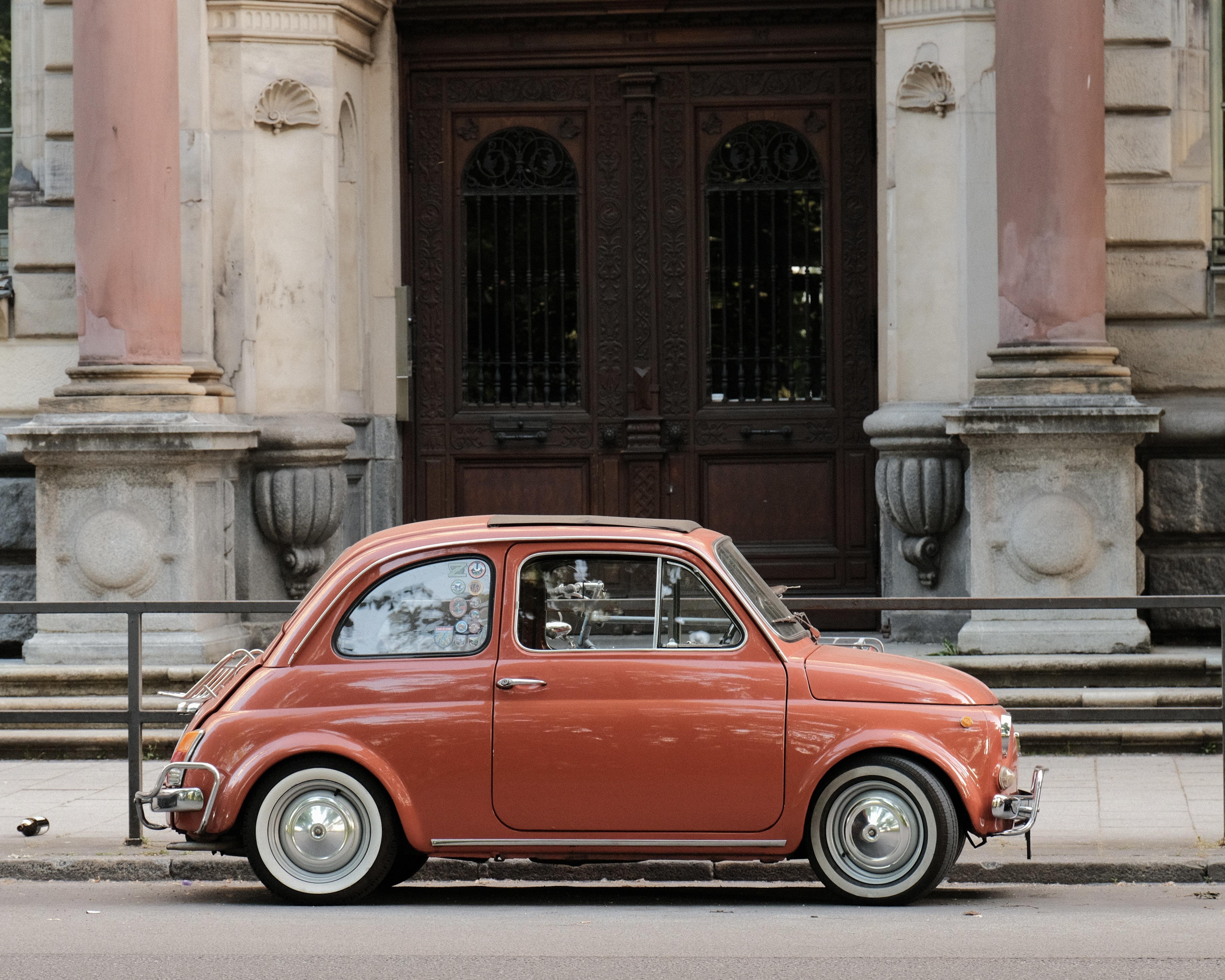 Side view of a small red vintage car with whitewall tires parked on a street in front of a stone building entrance with columns, steps, and dark wooden double doors.