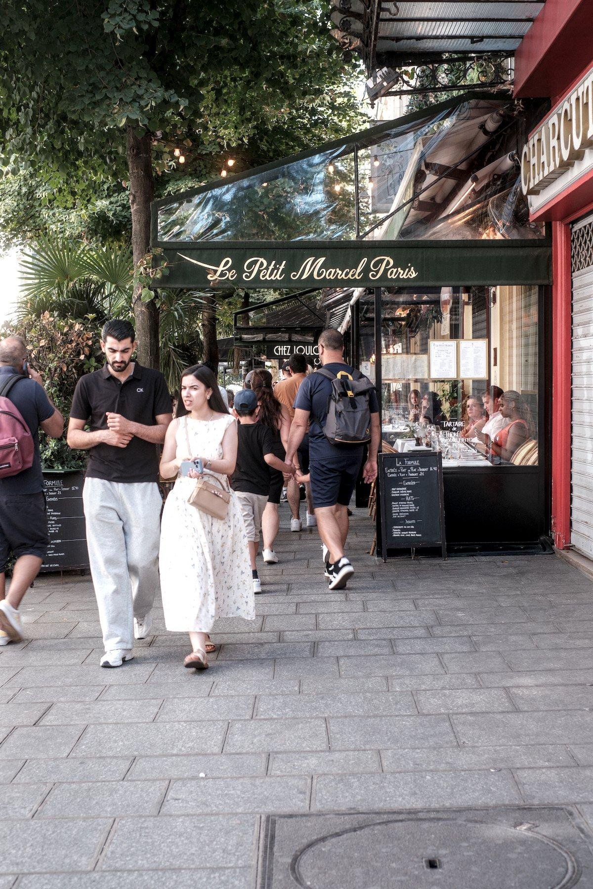 Photo verticale. Un trottoir parisien avec des terrace de cafés et leur paravent de délimitation. Des gens marchent dans les 2 sens. La première terrace est celle de « Le Ptit Marcel Paris » et plus loin on voit « Chez Loulou » dont le dernier U est caché par la tête d’un homme de dos.