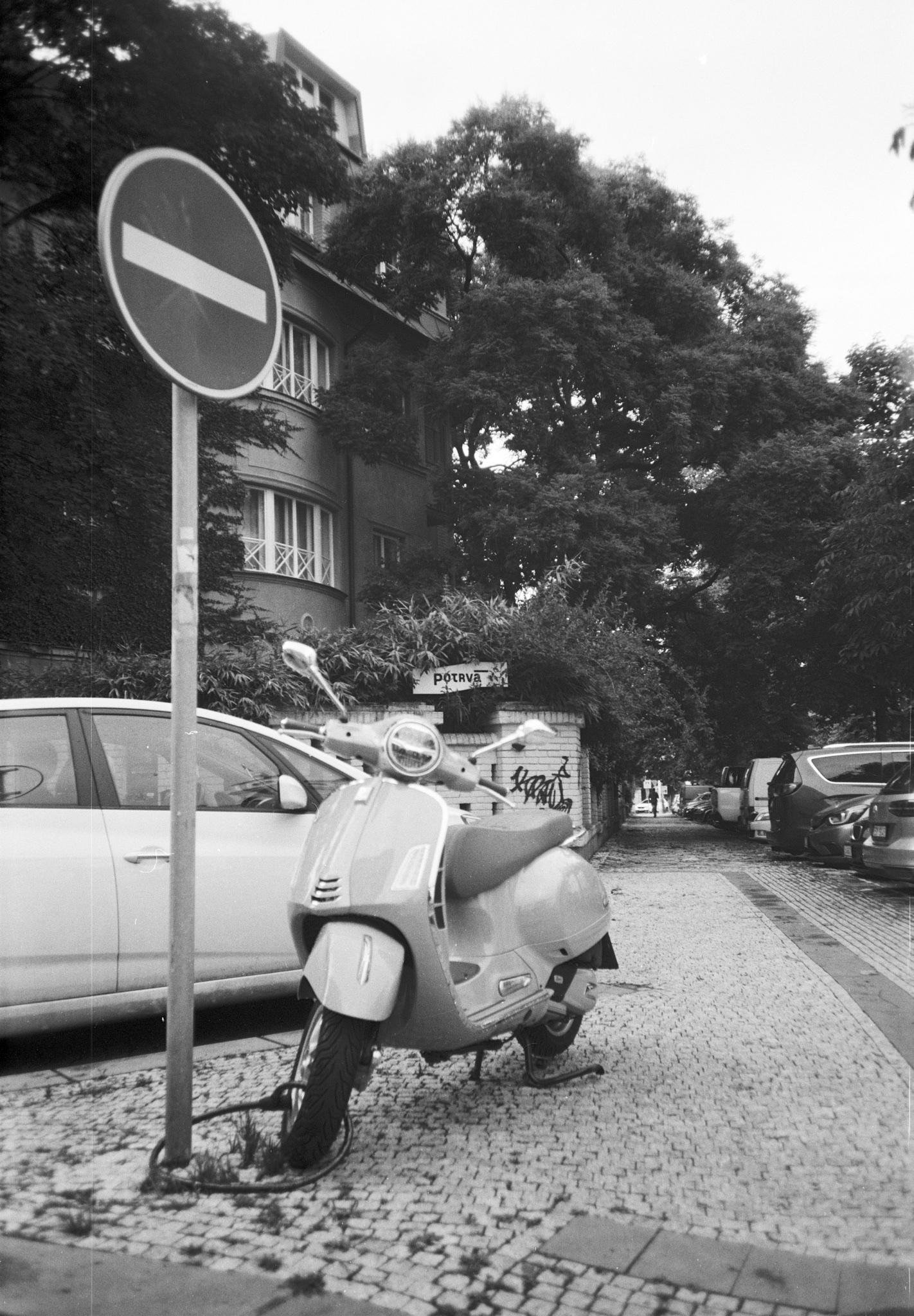 Black-and-white street scene with a “no entry” traffic sign in the left foreground and a parked scooter on a cobblestone sidewalk beside cars; a multi-story apartment building and large trees fill the background, with more parked vehicles lining the street to the right.