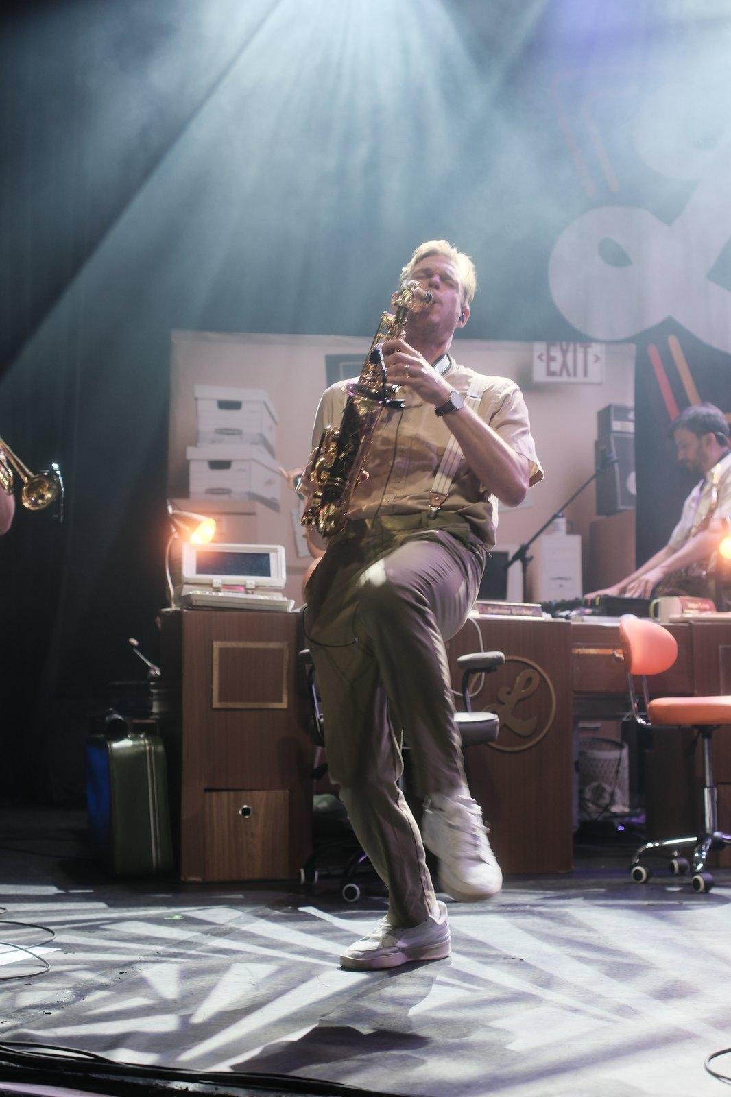 Photo verticale. Un musicien sur scène jouant du saxophone basculant en arrière, se tenant sur sa jambe droite et levant la gauche