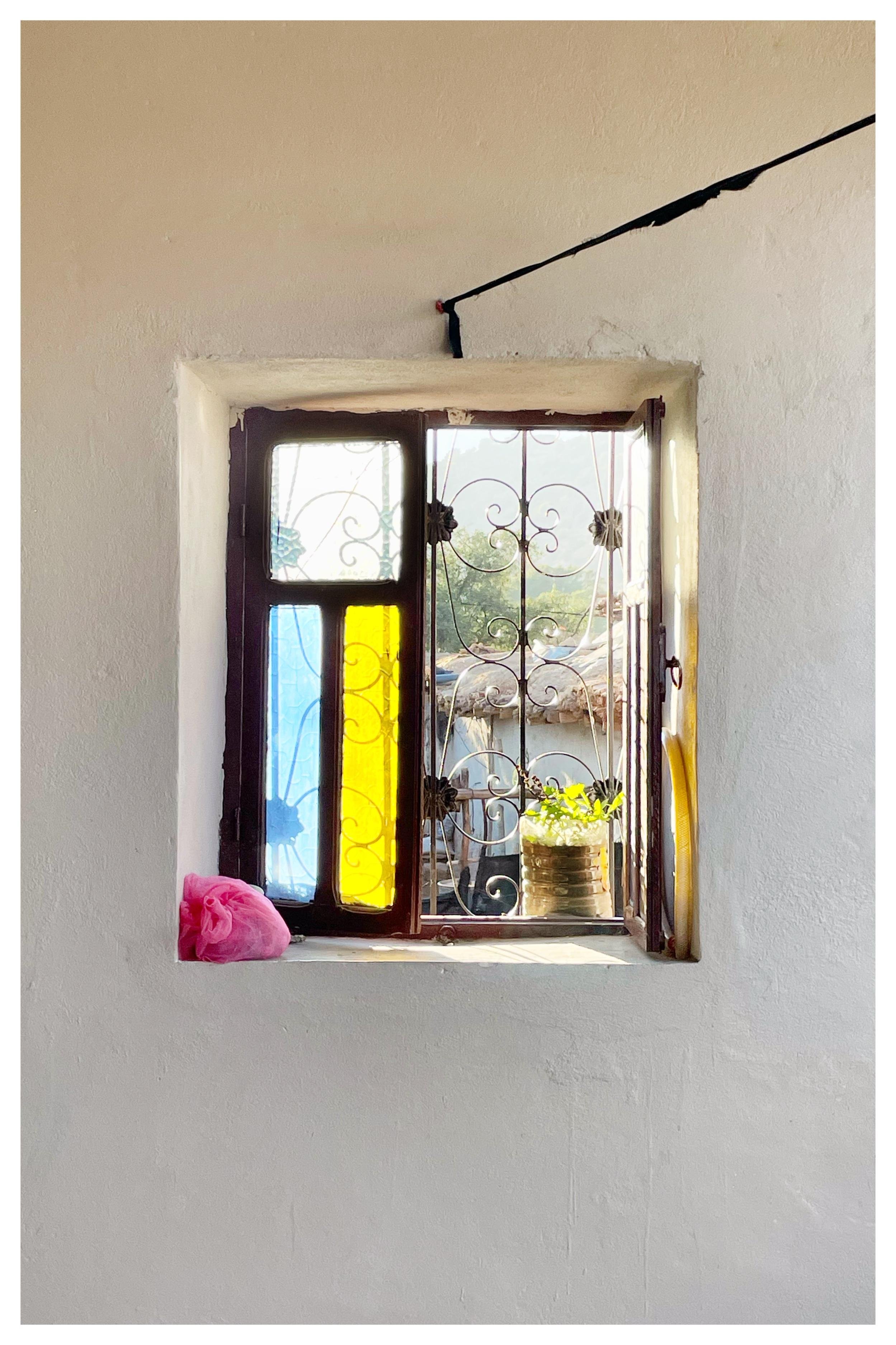 Small recessed window in a white wall with dark wooden frame and decorative metal grille; left pane has blue and yellow stained-glass sections, and the sill holds a pink cloth and a tin can planter with green leaves, with rooftops visible outside.