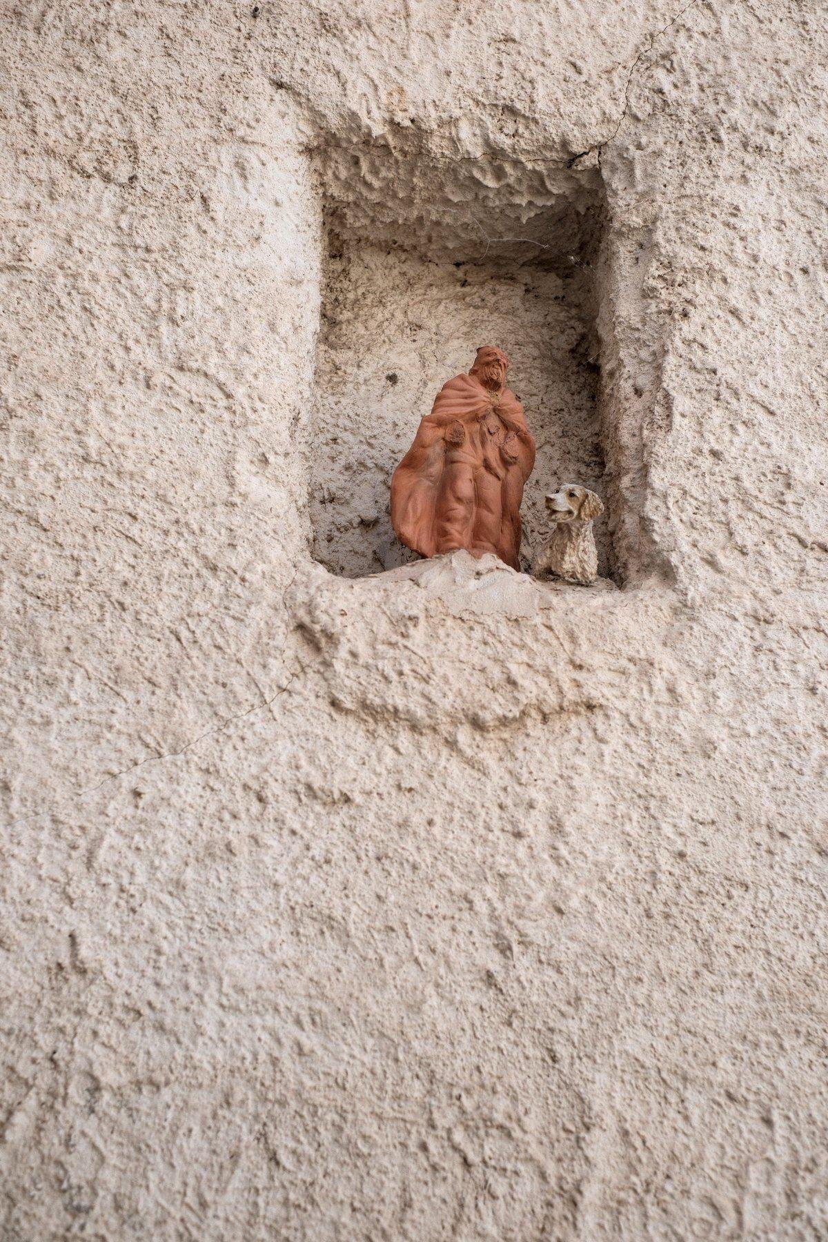 Photo verticale. Dans un mur crépifié, une cavité rectangulaire dans laquelle se trouve une figurine d’un vieil homme et d’un chien