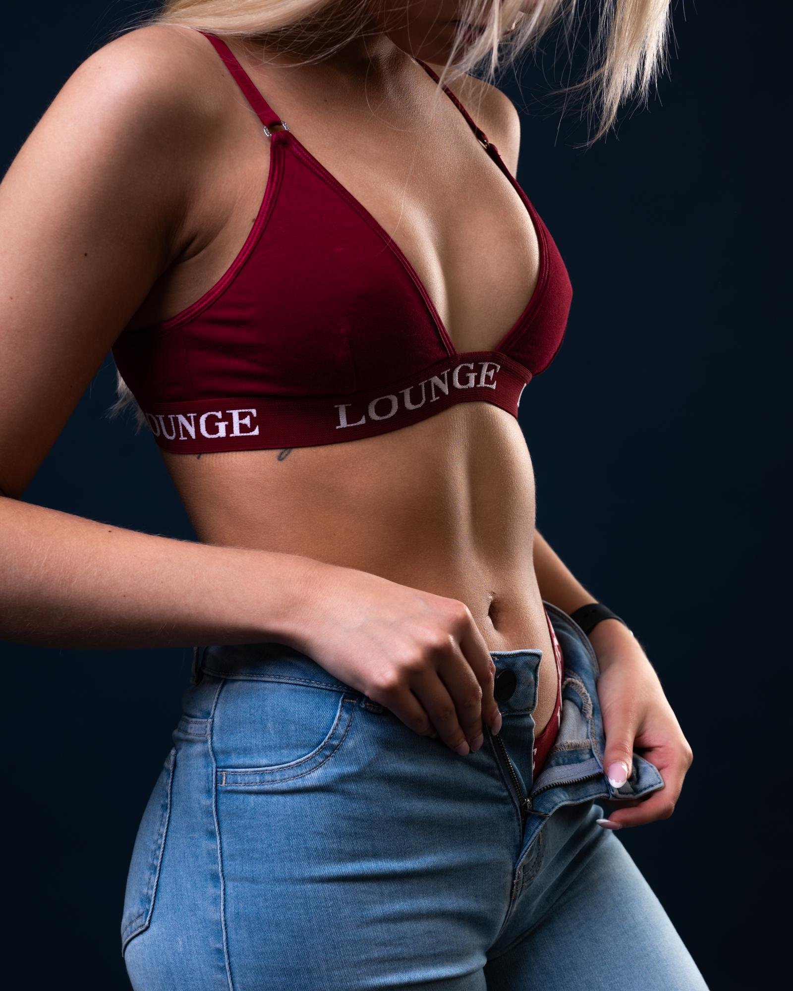 Cropped studio photo of a woman from shoulders to hips wearing a burgundy bra with “LOUNGE” on the band and blue jeans that are unbuttoned and partially unzipped, hands holding the waistband against a dark background.